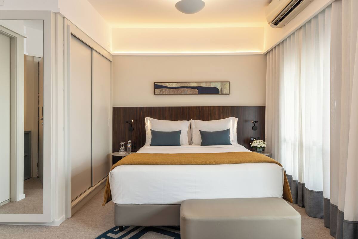 Quarto de hotel moderno com uma cama grande, uma mesa de cabeceira com abajur e vista para a cidade pela janela.