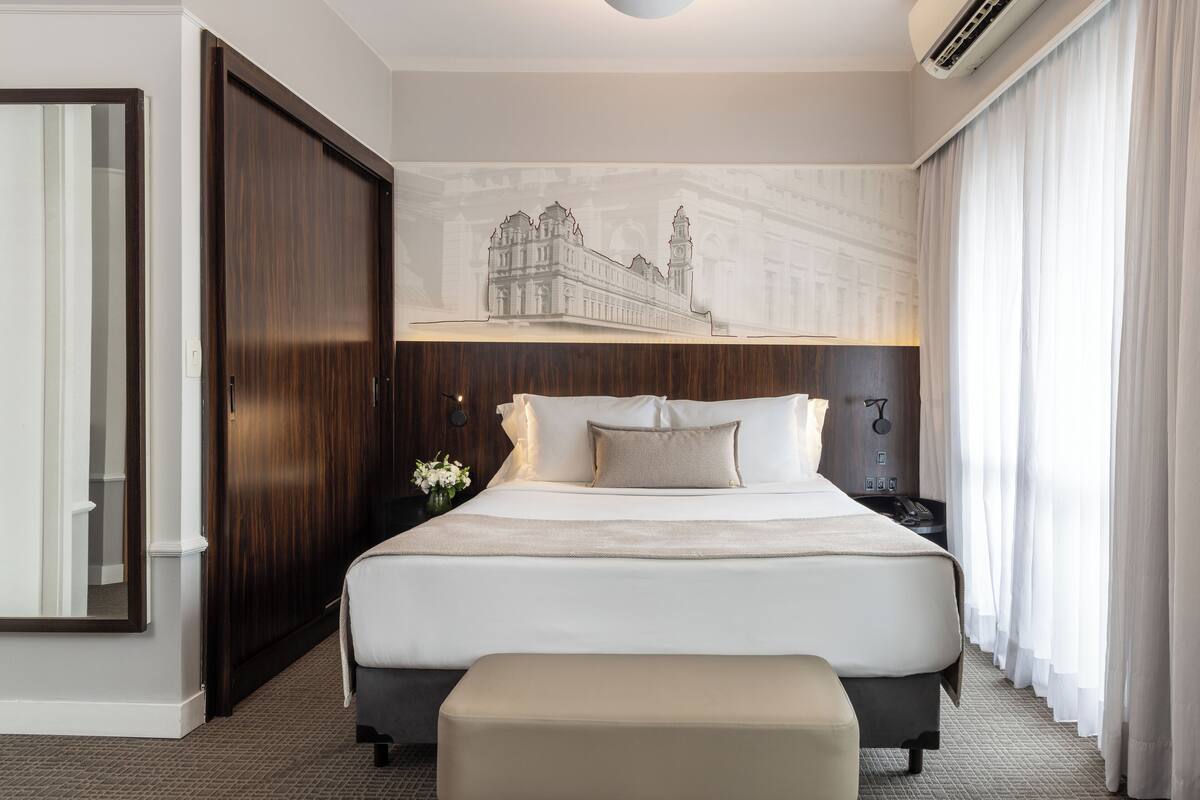 Quarto de hotel com uma cama grande, mesa de cabeceira, luminária na parede e um quadro de um edifício.