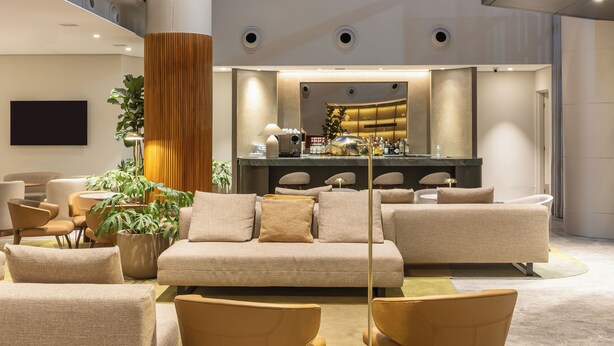Un vestíbulo amplio con techo alto, zona de estar central con sofás y sillones, área de bar y un gran ventanal que proporciona luz natural.