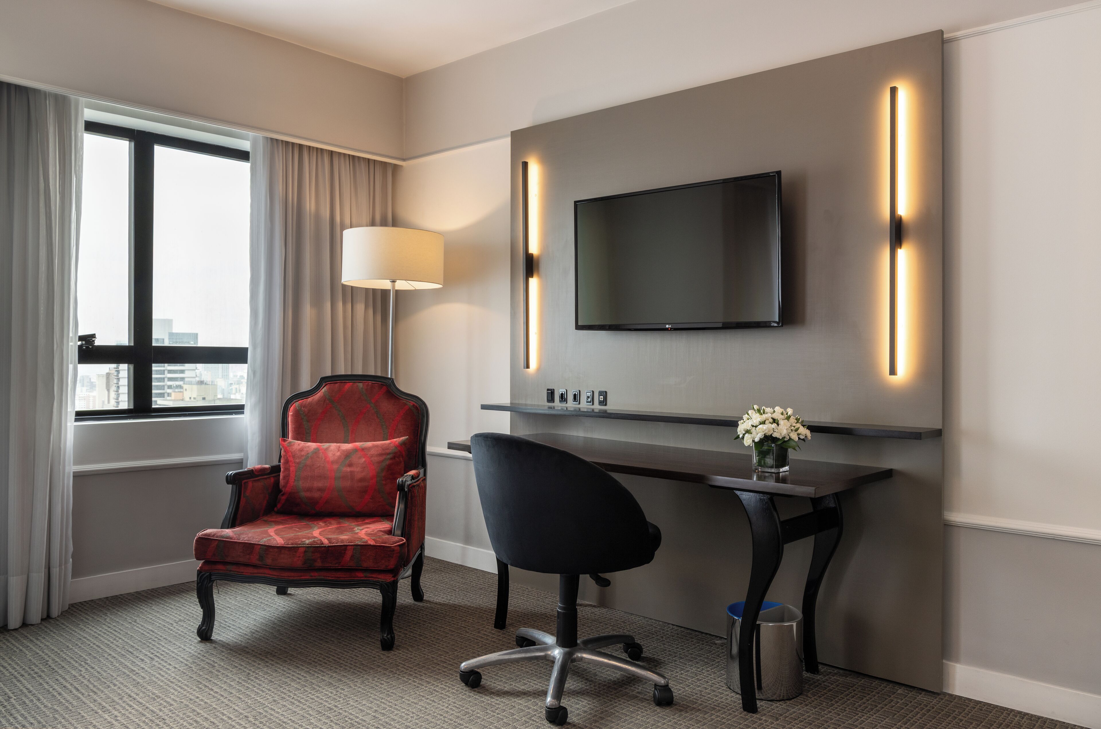 Ein Hotelzimmer mit großem Fenster, einem roten Polsterstuhl, einem schwarzen Bürostuhl, einem Schreibtisch mit Flachbildfernseher und einer Vase mit Blumen.