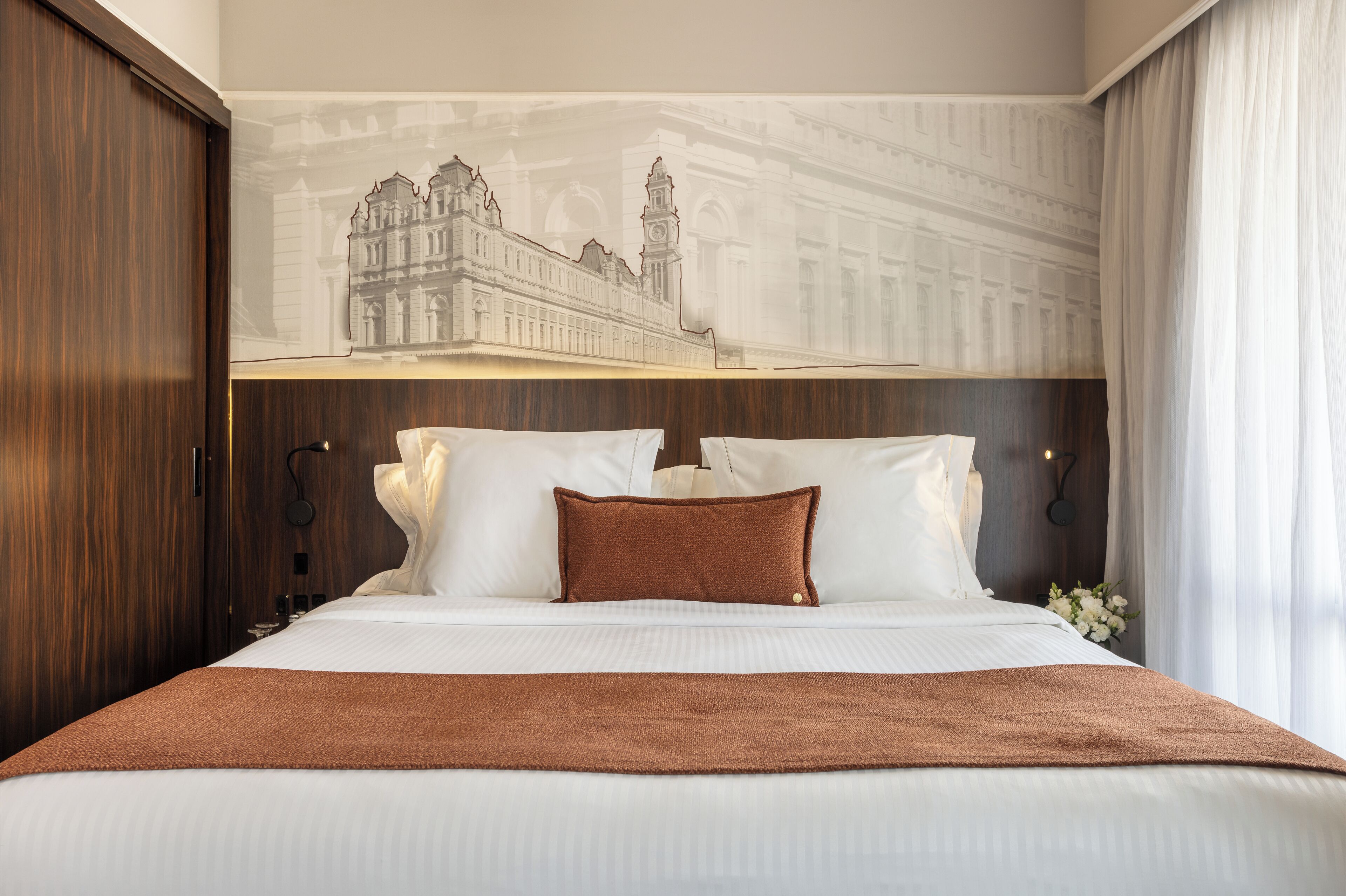 A hotel room with a large bed, a headboard with a cityscape mural, a wooden wardrobe, and a nightstand with a lamp.