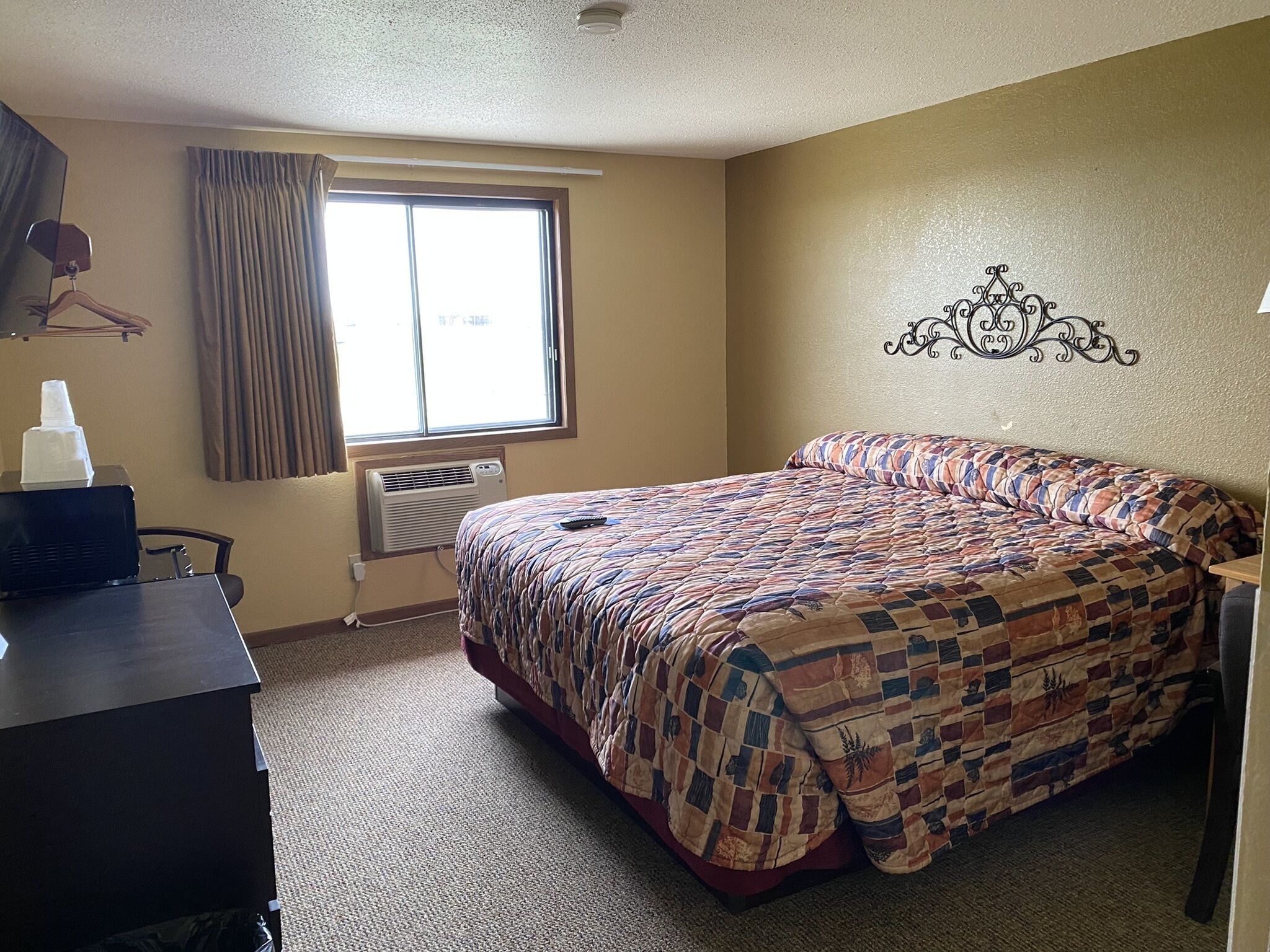 Habitación de hotel con cama, escritorio, silla, ventana con cortinas, aire acondicionado y un adorno en la pared.