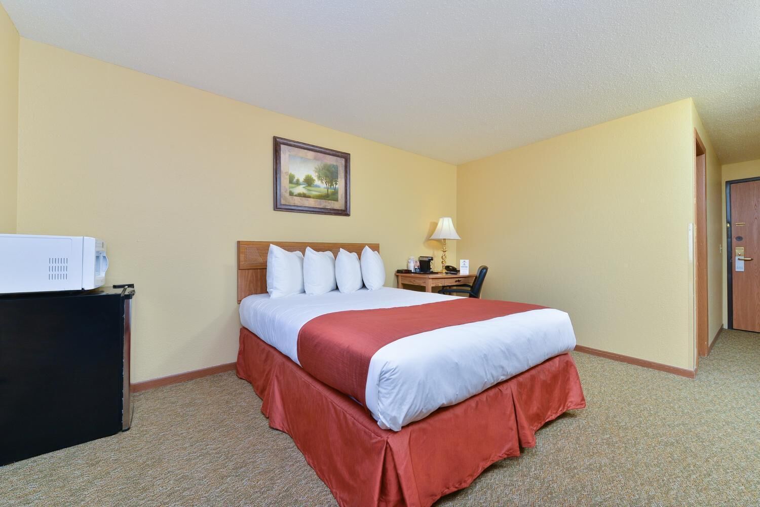 A hotel room with a bed, a desk with a lamp, a microwave, and a painting on the wall.