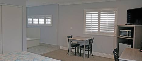 A bedroom with a bed, a desk, and a chair. There is a window with blinds, a microwave, and a television.