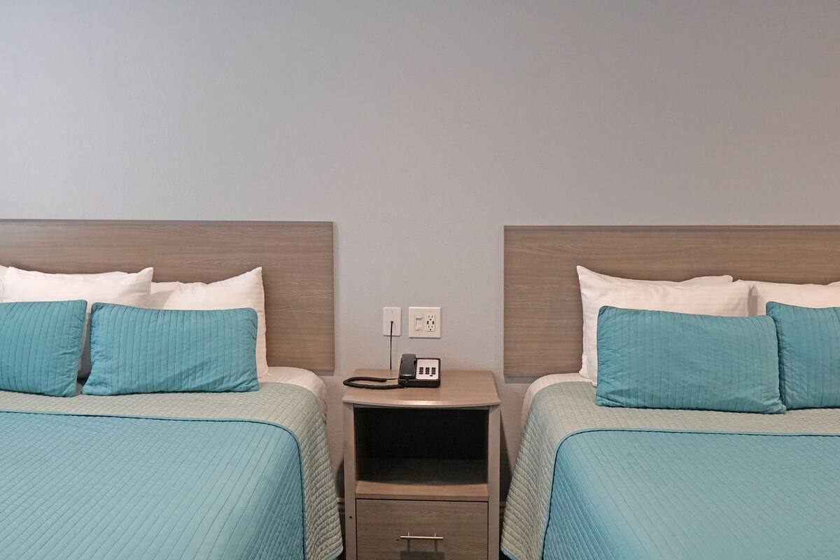 A hotel room with two beds, each with a teal bedspread and white pillows. There is a bedside table with a phone and a power outlet.