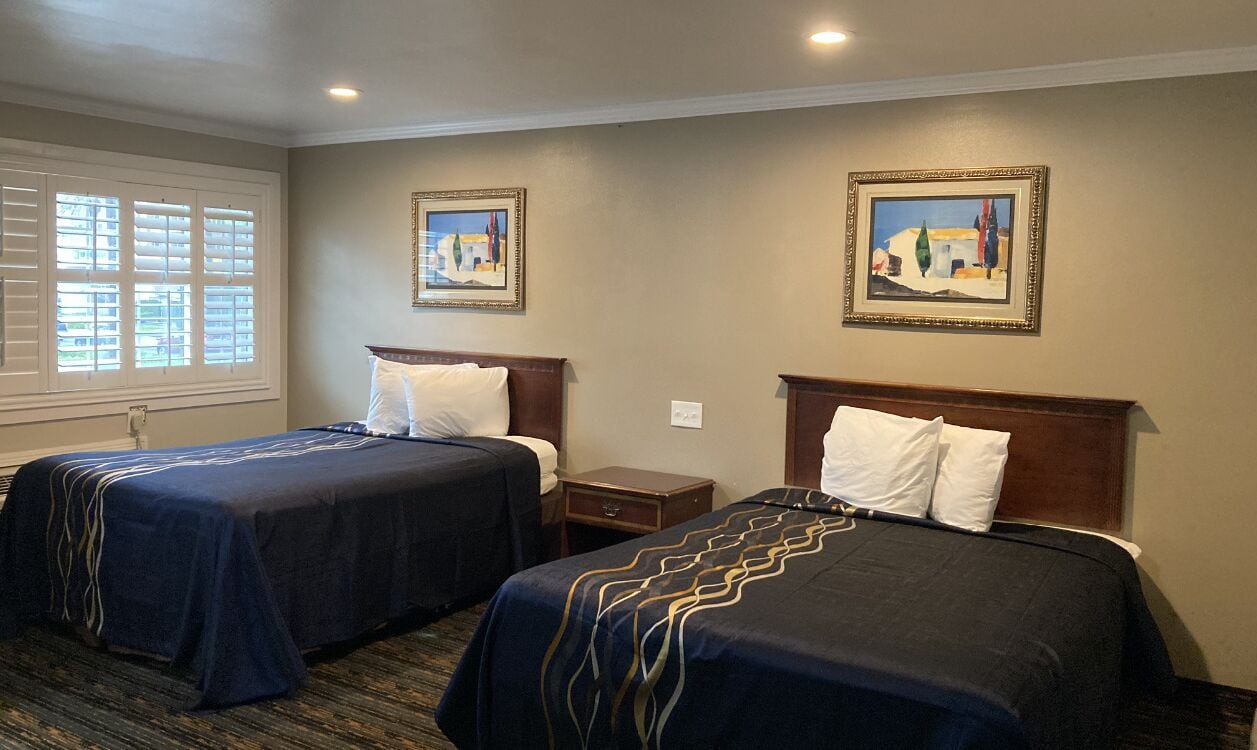 A hotel room with two beds, a window with blinds, a nightstand, and two framed pictures on the wall.