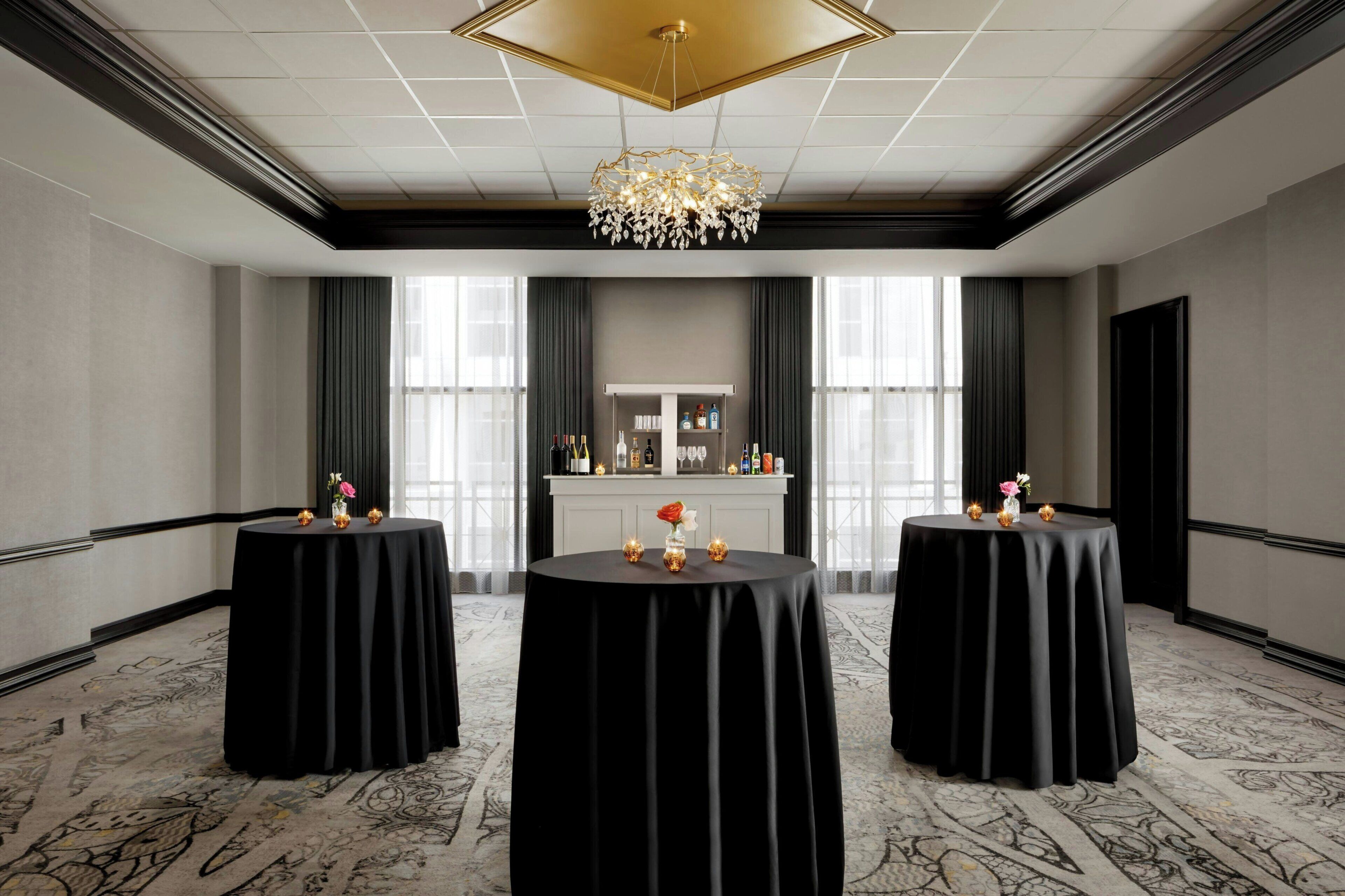 Une chambre d’hôtel avec un lustre, deux tables rondes recouvertes de nappes noires, une cheminée et une vue dégagée sur l’extérieur grâce à de grandes fenêtres.