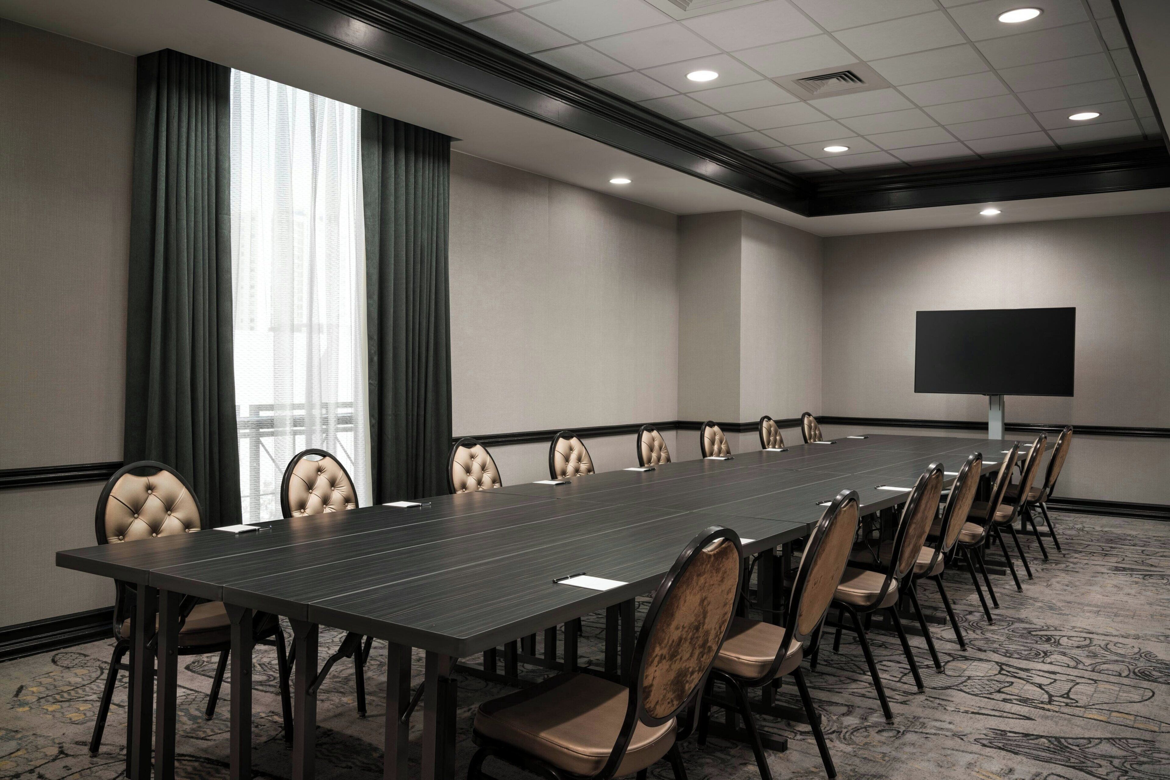 A conference room with a long table, chairs, a projector screen, and a view of buildings through large windows.