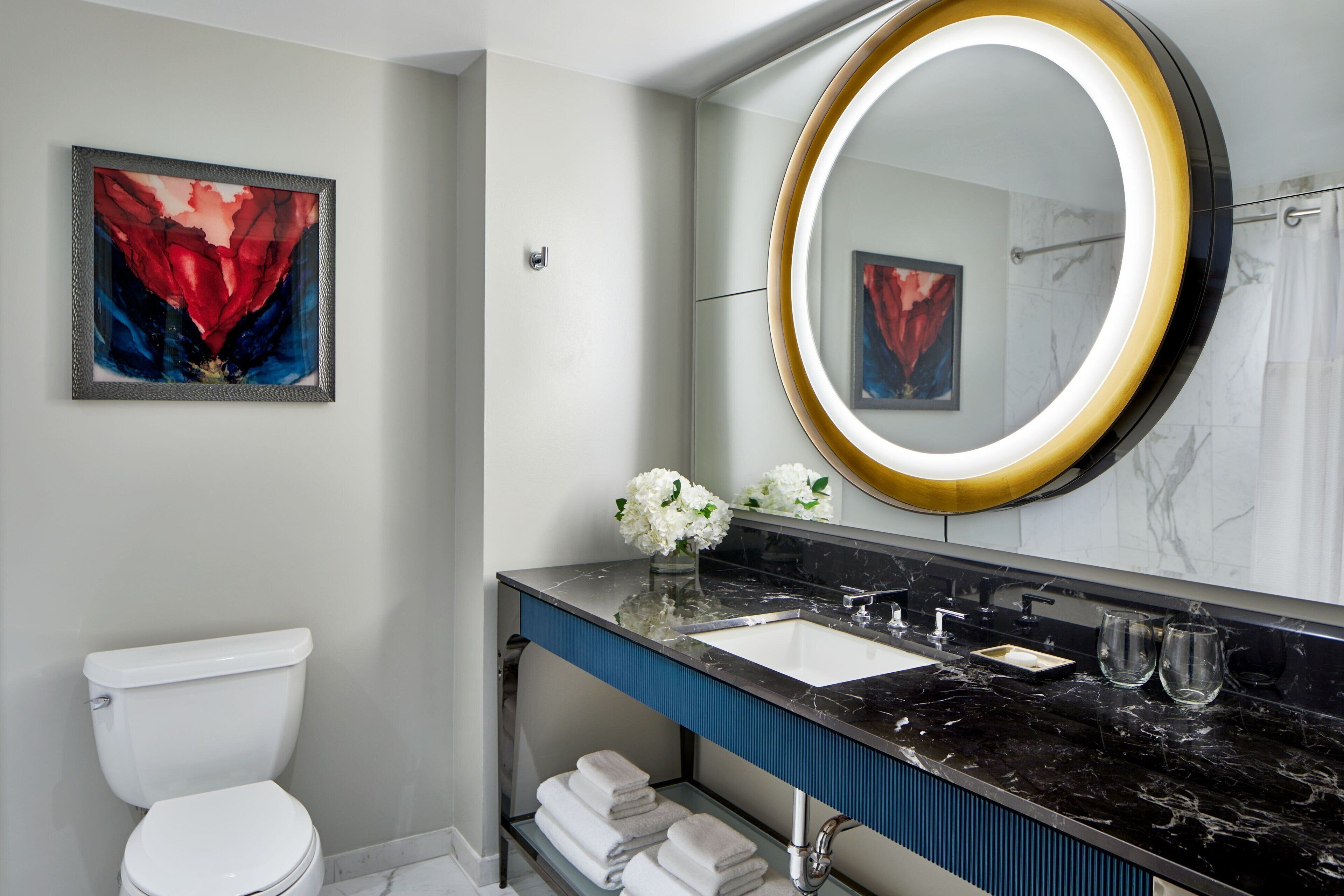 A modern bathroom with a white toilet, a black marble sink, a large mirror with a gold frame, and a framed abstract painting on the wall.