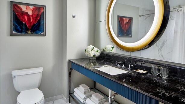 A modern bathroom with a white toilet, a black marble sink, a large mirror with a gold frame, and a framed abstract painting on the wall. - Hotel Beauden, Autograph Collection (Orlando)