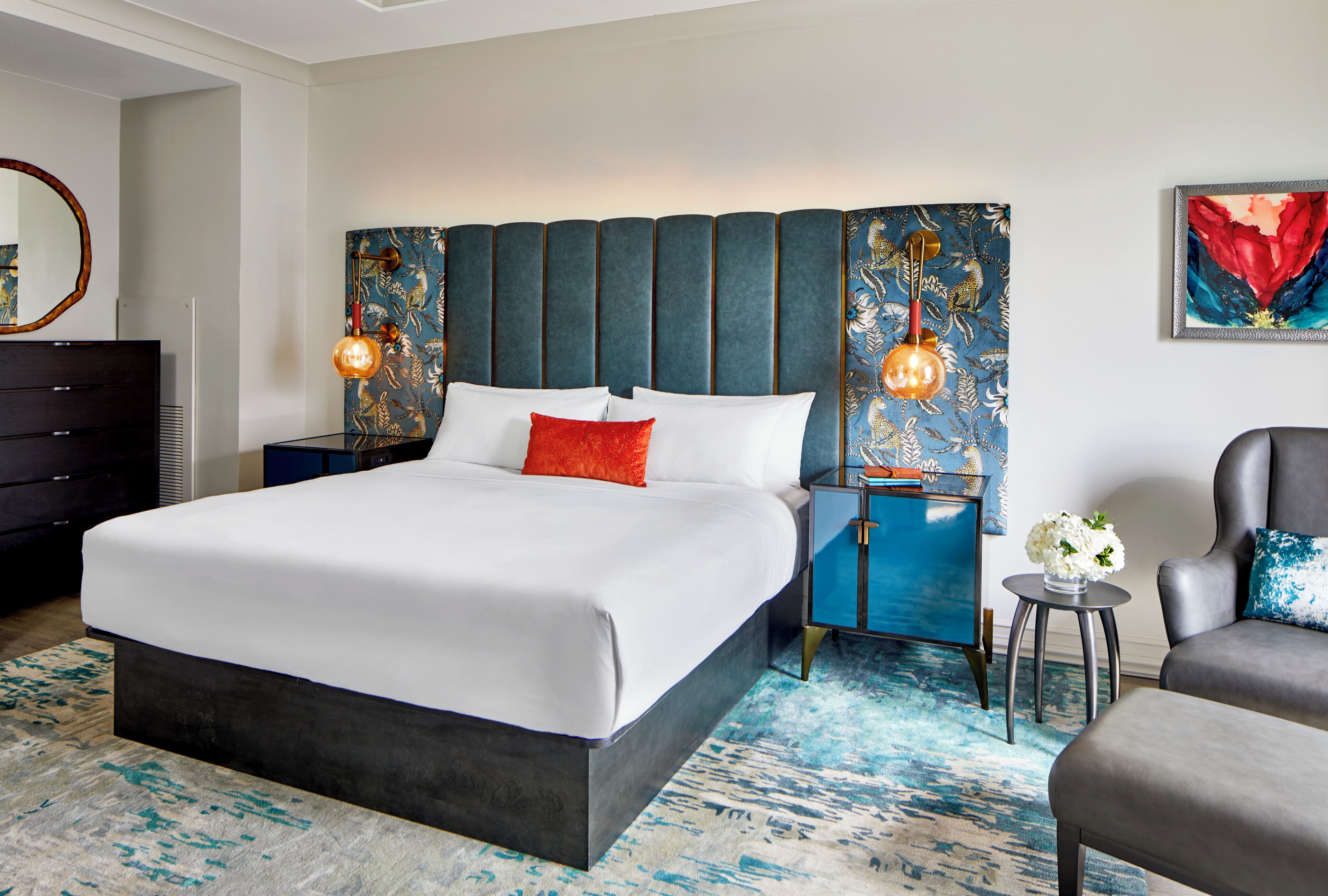 Una habitación de hotel moderna con una cama grande, cabecera con paneles verticales, mesita de noche con lámpara, una silla y una pequeña mesa con flores.