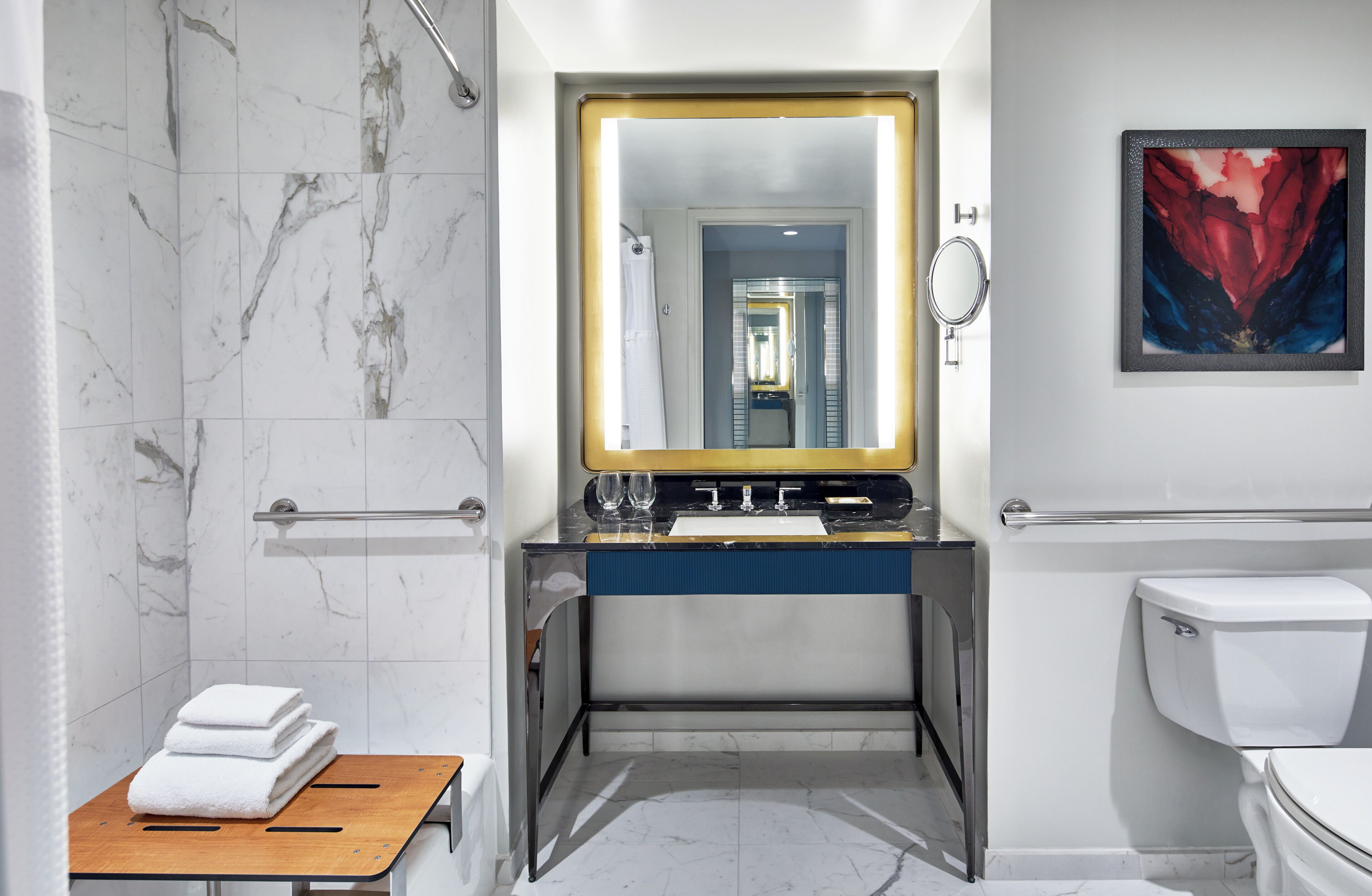 A bathroom with a marble shower, a sink with a gold mirror, a toilet, and a framed artwork on the wall.