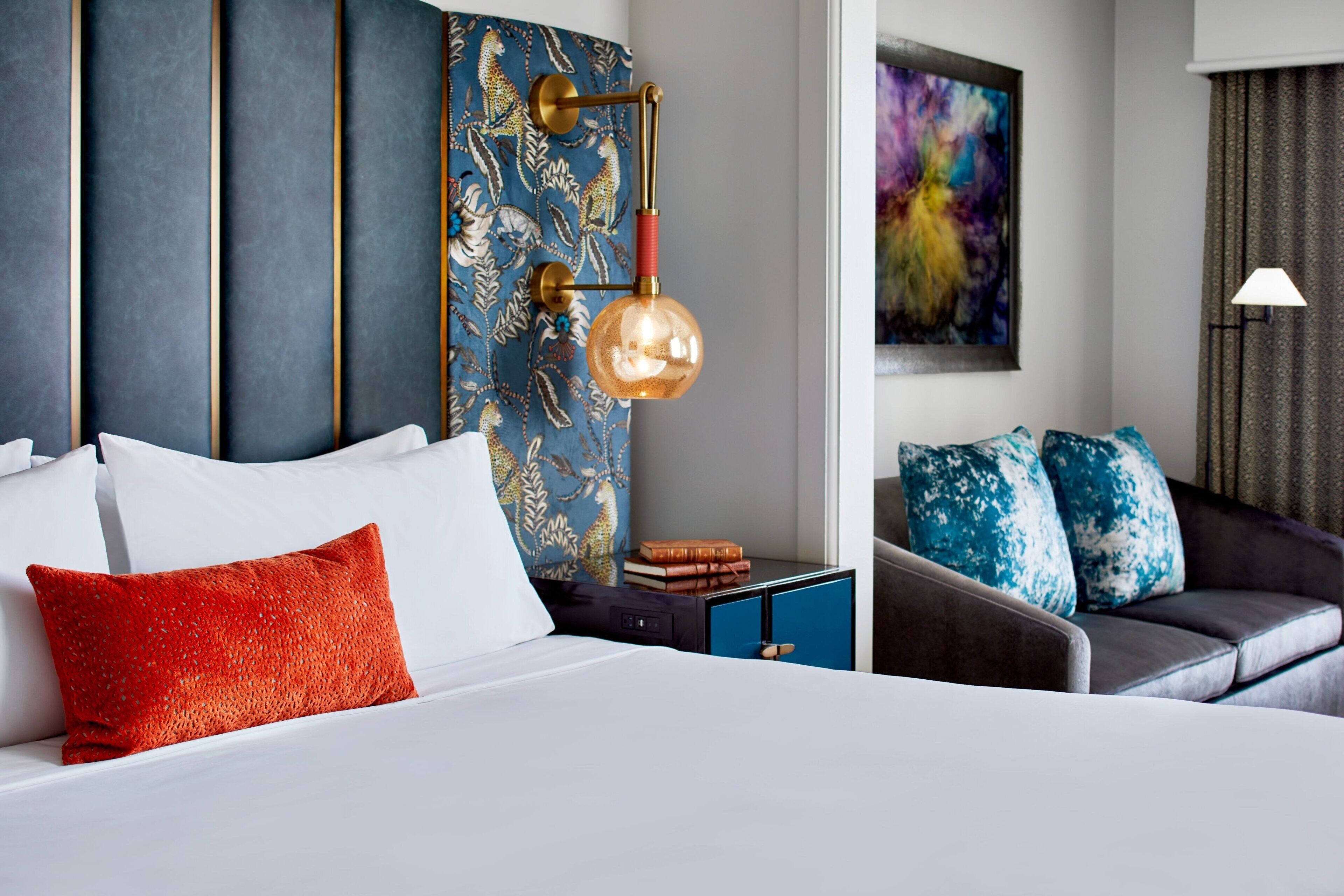 A modern hotel room with a bed, a nightstand, a blue cabinet, a sofa, and a wall-mounted lamp.