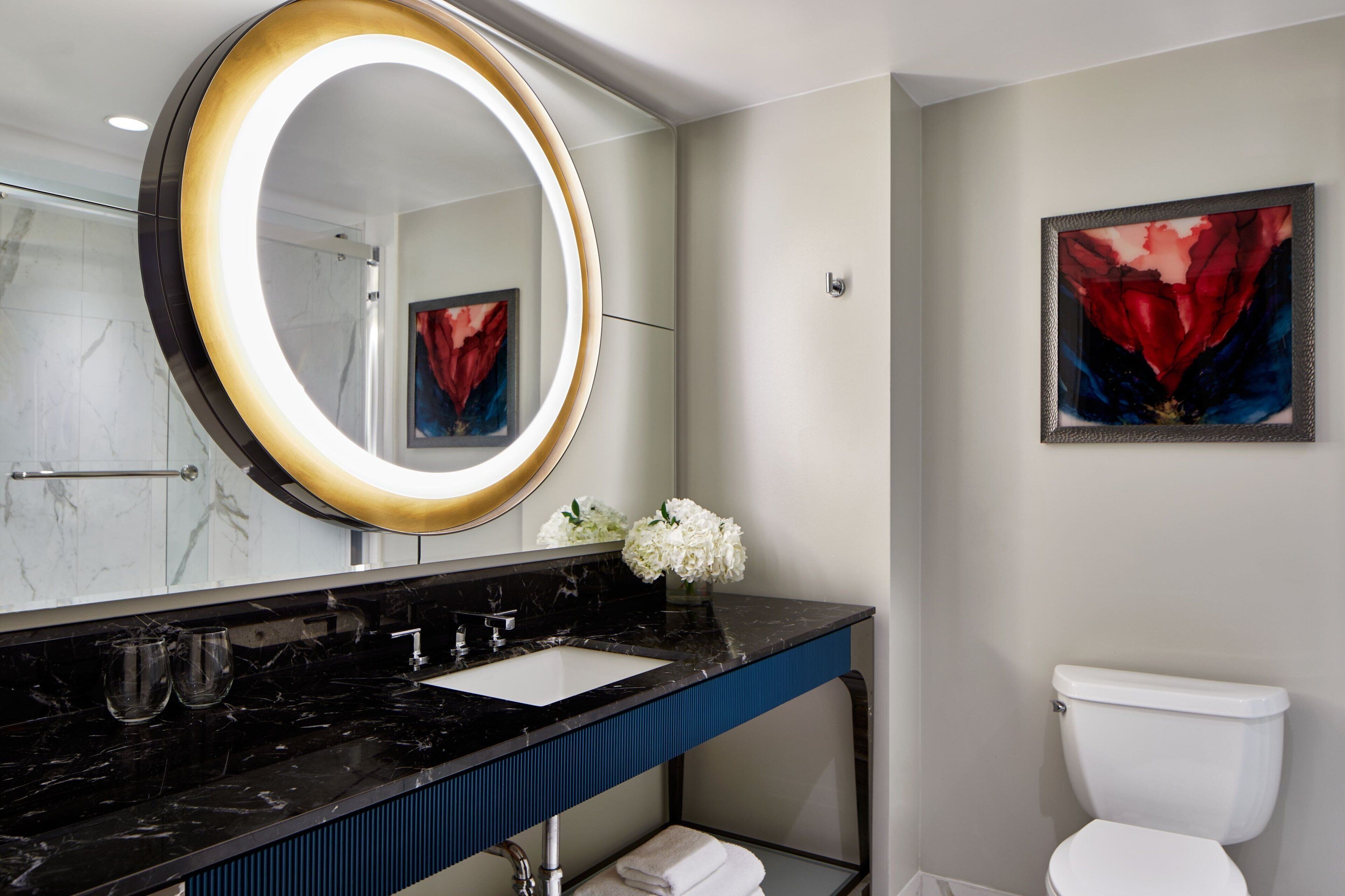 A modern bathroom with a black marble sink, a round mirror with a light, a white toilet, and a framed abstract painting on the wall.