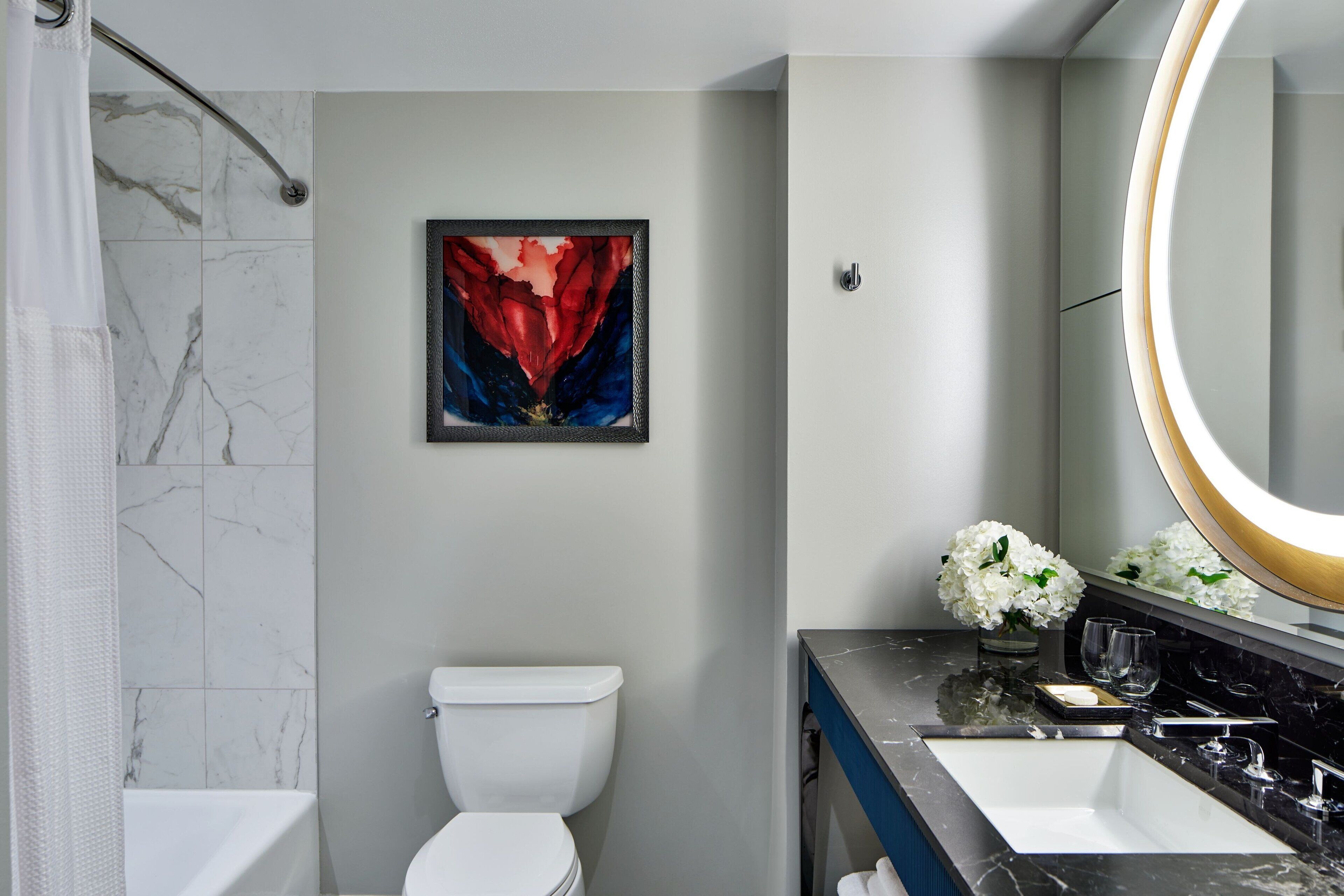 Un baño moderno con inodoro blanco, zona de ducha con azulejos de mármol, un espejo grande con marco dorado y un cuadro abstracto enmarcado.