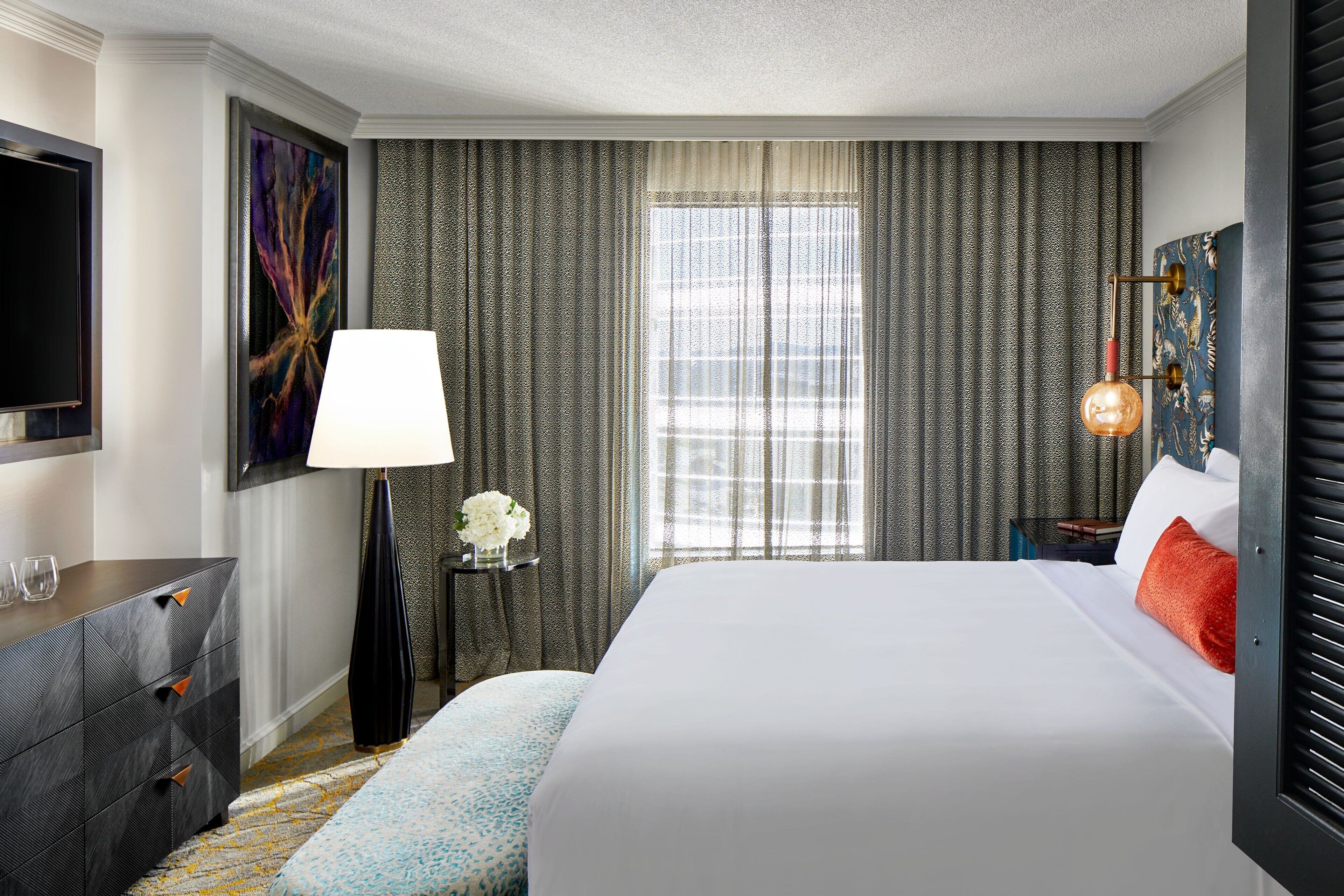 A hotel room with a large bed, a flat-screen TV, a desk with drawers, a lamp, and a window with curtains.