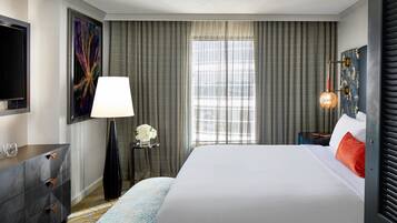 A hotel room with a large bed, a flat-screen TV, a desk with drawers, a lamp, and a window with curtains.