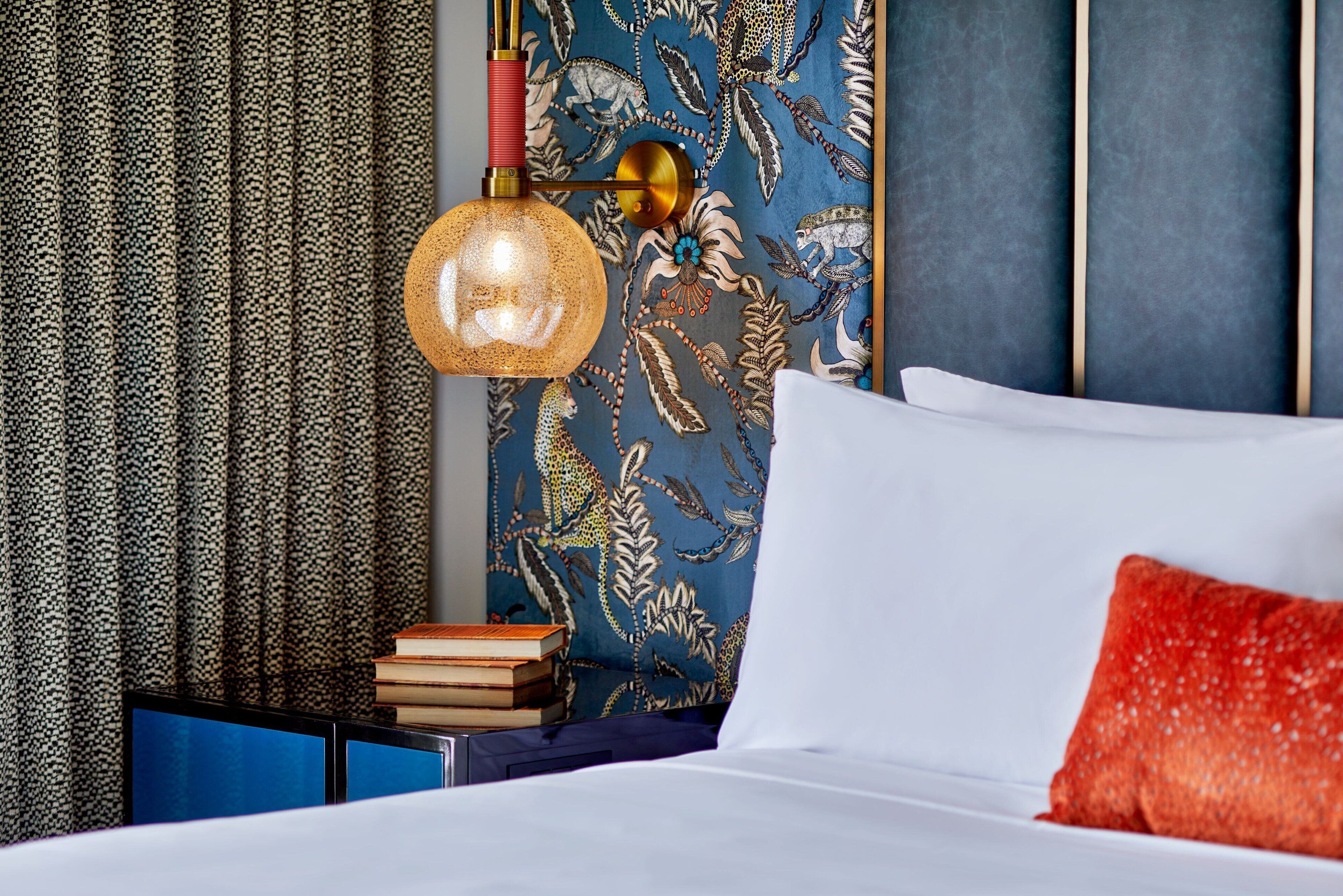 Een net opgemaakt bed met een wit en rood kussen, een stapel boeken op een blauwe nachtkastje, en een muur met een blauw bloemendesign.
