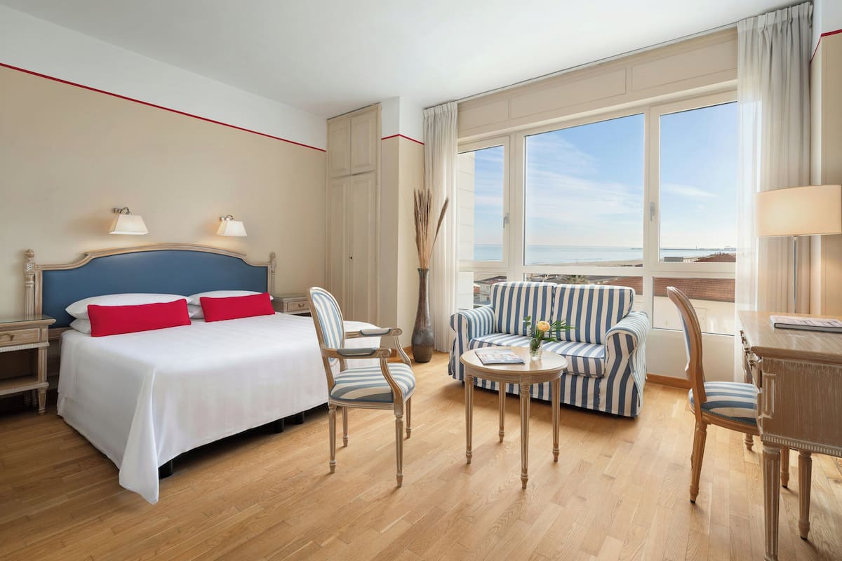 A hotel room with a bed, two chairs, a small table, a sofa, and a view of the sea.