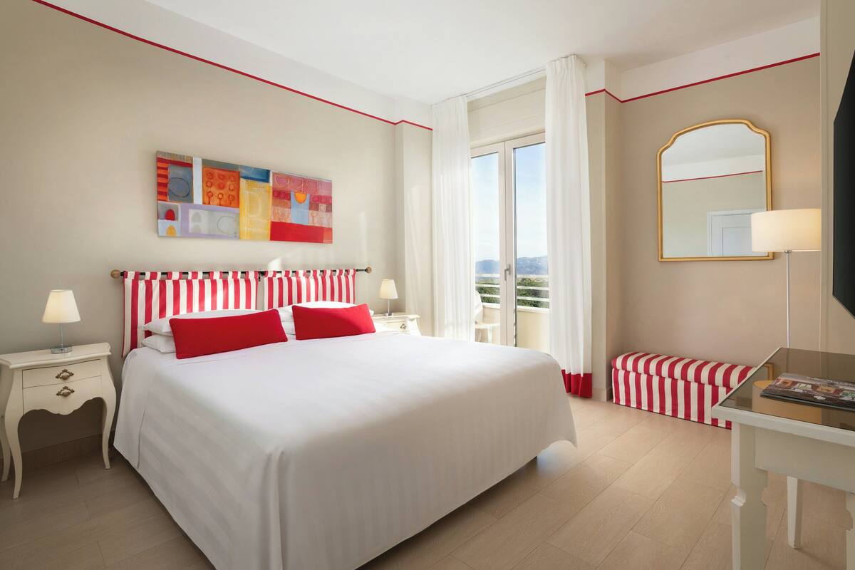 A hotel room with a large bed, a bedside table with a lamp, a small bench, a large window with curtains, and a wall-mounted mirror.