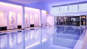 Un área de piscina interior moderna con paredes de vidrio transparente, una piscina central y estatuas iluminadas sobre pedestales.