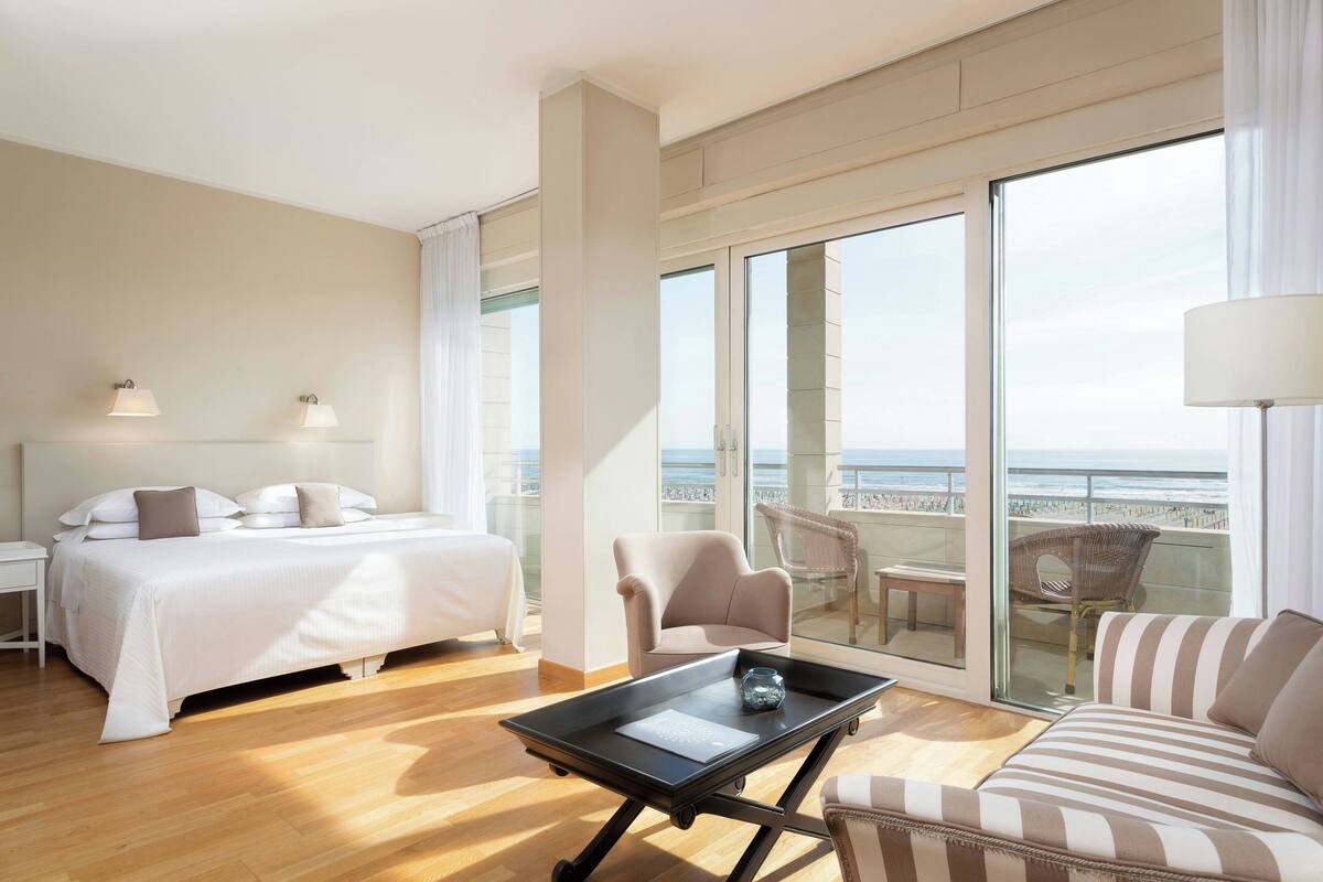 A hotel room with a bed, a sofa, a table, a chair, and a balcony with a view of the beach.