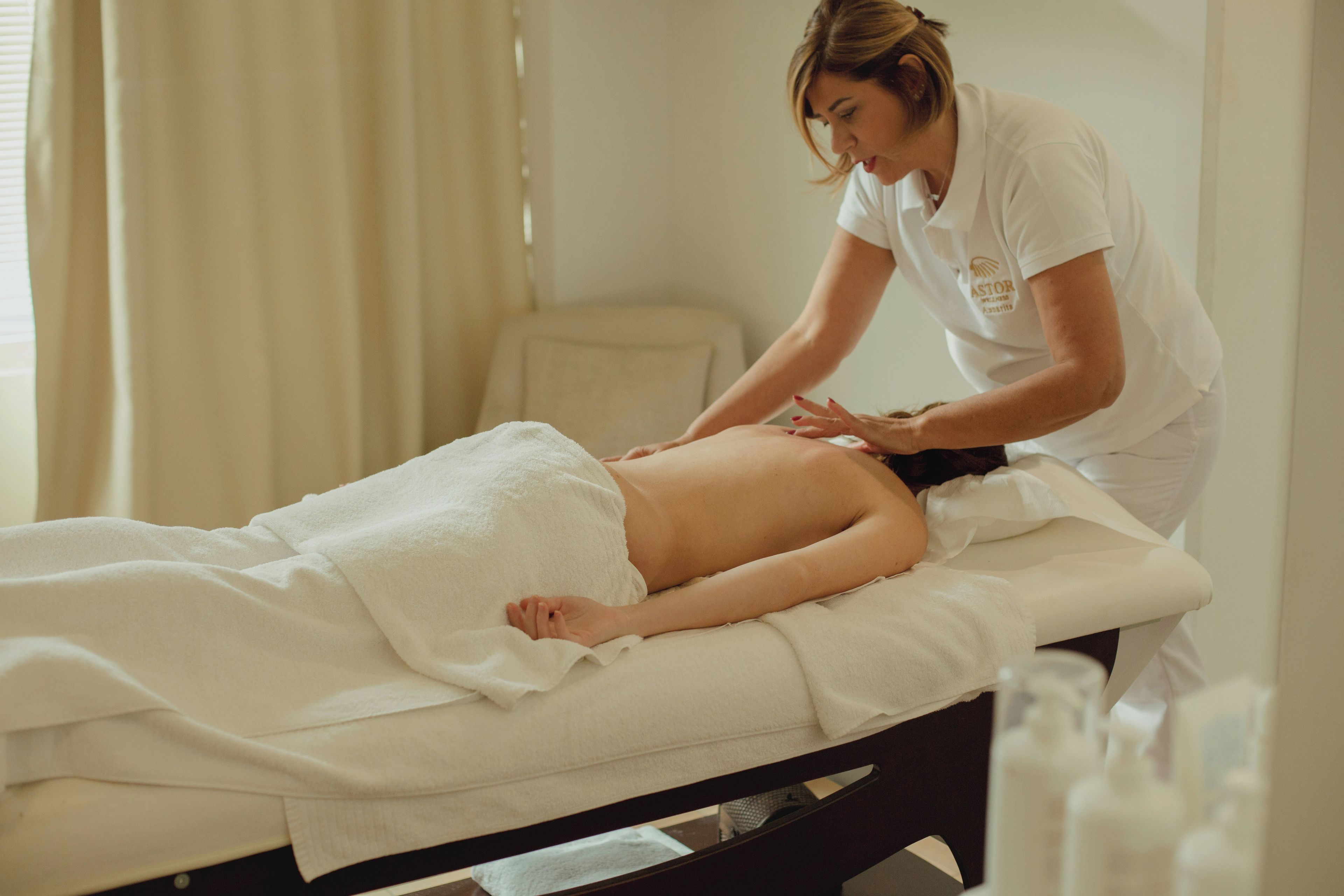 A massage therapy session in a room with a bed, a chair, and a window with curtains.