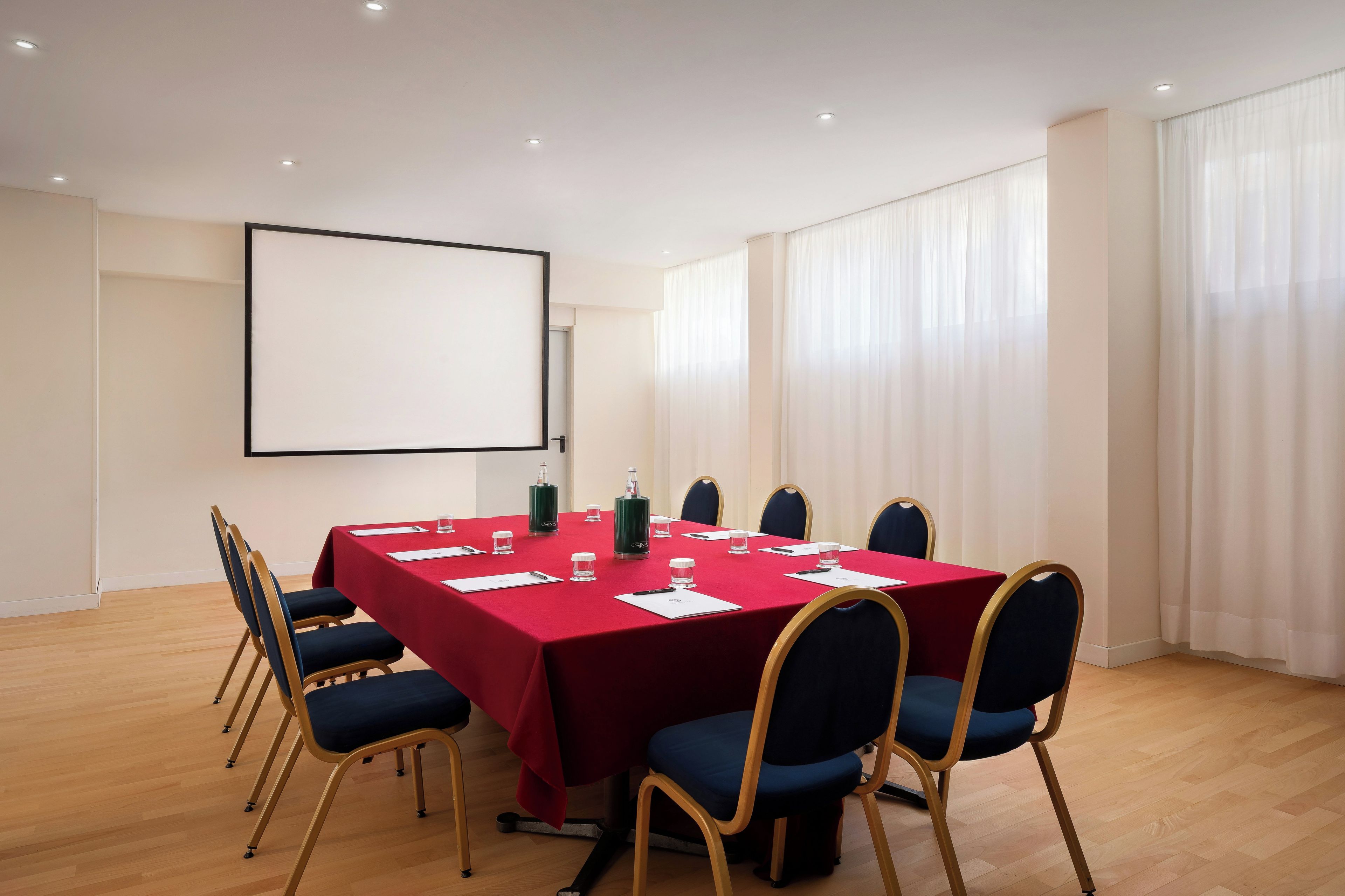 A conference room with a red table, blue chairs, a projector screen, and large windows with curtains.