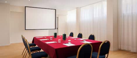 A conference room with a red table, blue chairs, a projector screen, and large windows with curtains.