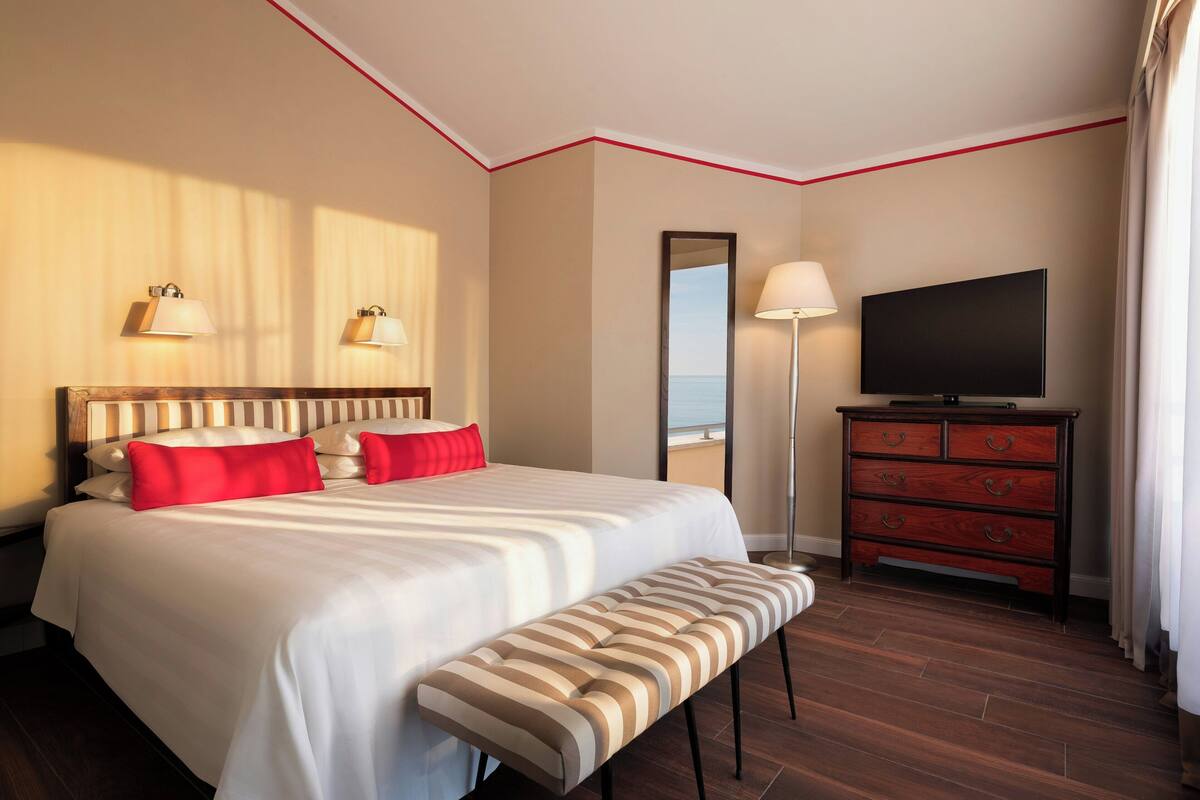 A hotel room with a large bed, bedside lamps, a flat-screen TV, a dresser, and a bench.