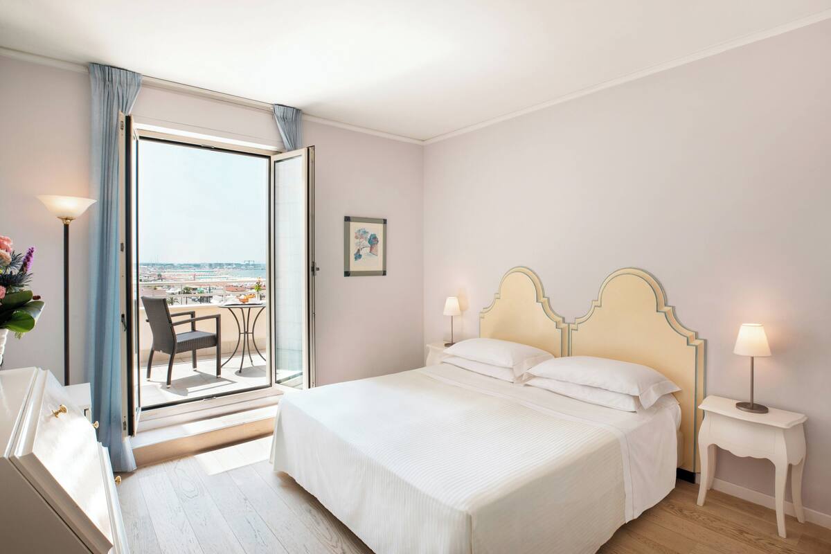 A hotel room with a bed, a nightstand, a lamp, a picture on the wall, and a view of the city through a glass door.