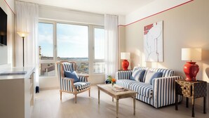 Una sala de estar moderna con sofá, sillón y mesa de centro, una lámpara roja y vista a la ciudad a través de amplios ventanales.