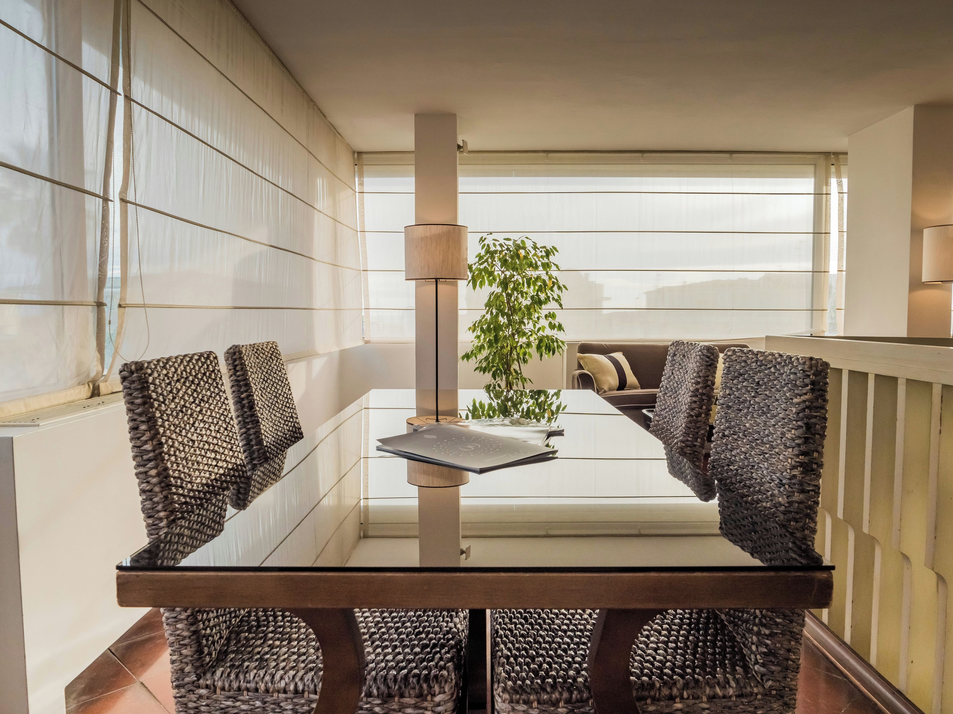 Een moderne eetruimte met een glazen tafel, gevlochten stoelen, een potplant en uitzicht door een raam.