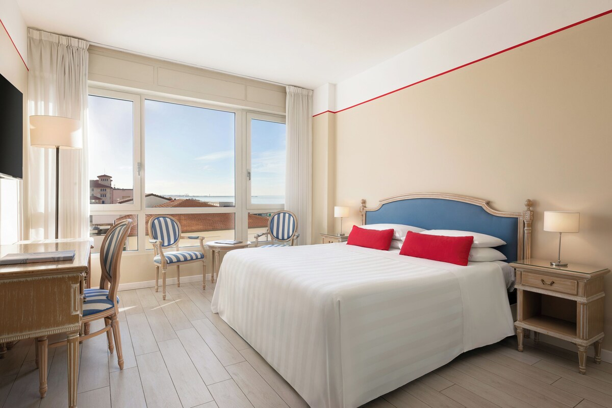 A hotel room with a large bed, a desk, two chairs, a nightstand, and a TV.