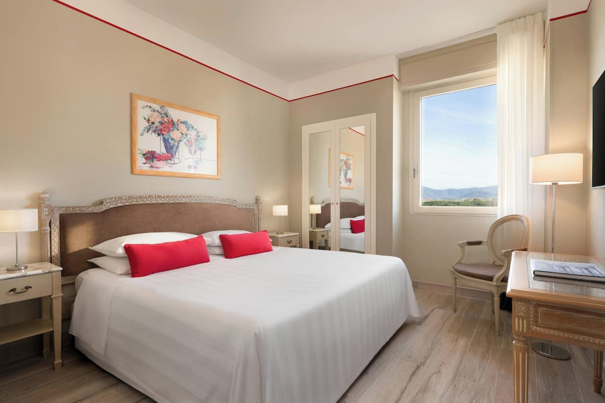 A hotel room with a large bed, a desk, a chair, a nightstand, a lamp, a painting, a mirror, and a window with a view.