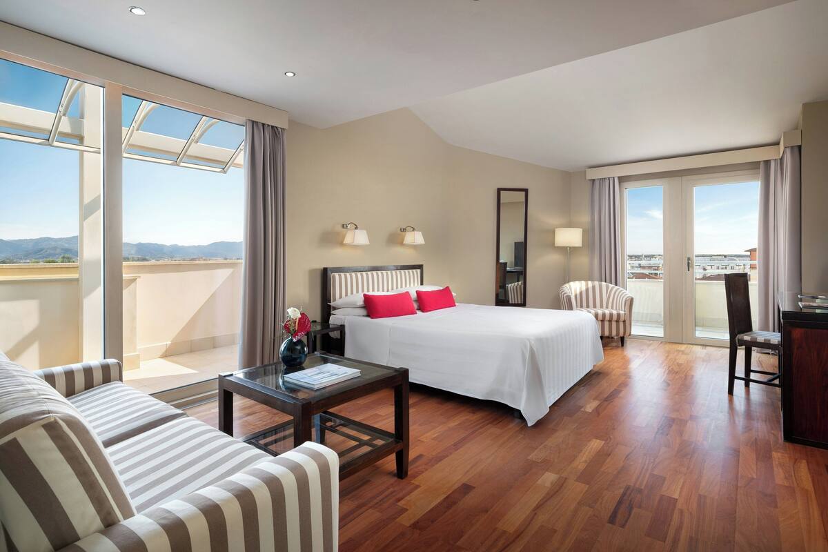 A hotel room with a bed, a sofa, a desk, a chair, and a balcony with a view.