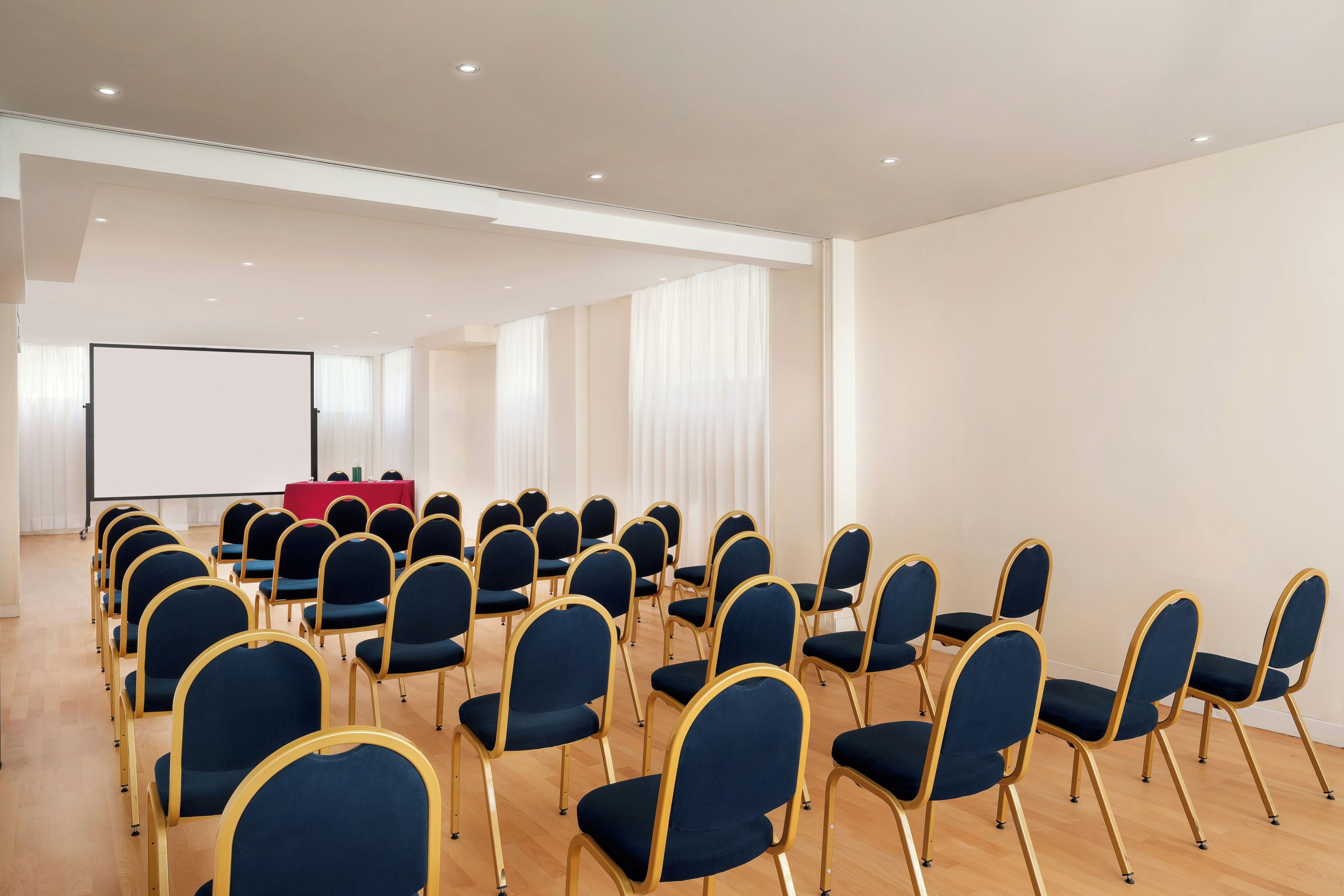 A conference room with rows of chairs, a projection screen, and a red table.