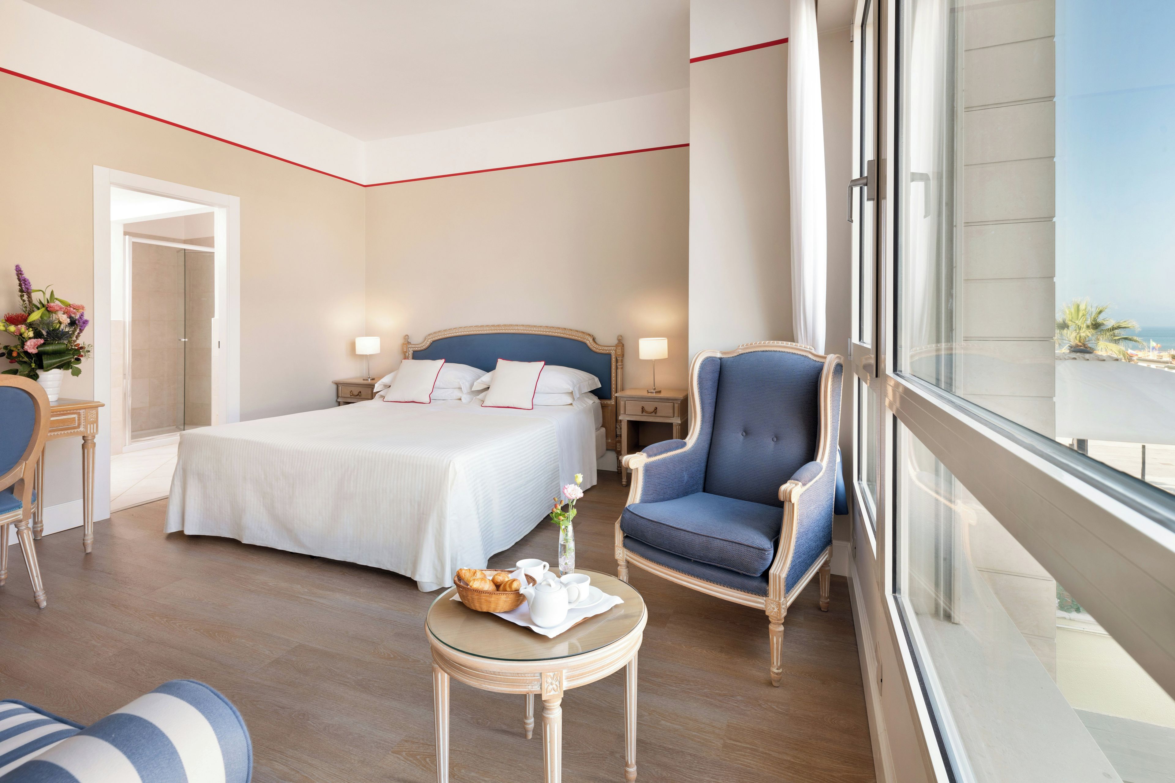 Une chambre d’hôtel avec un grand lit, un coin salon comprenant un fauteuil et une petite table, une vue sur la mer par la fenêtre, et une porte donnant sur une autre pièce.