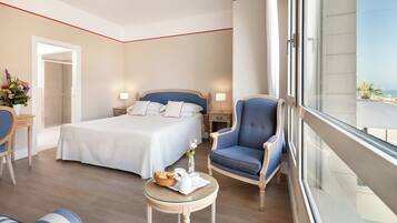 Una camera d'albergo con un letto grande, un angolo salotto con una poltrona e un tavolino, vista mare attraverso una finestra e una porta che conduce a un'altra stanza.