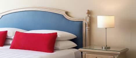 Una cama bien hecha con almohadones blancos y rojos, un cabecero azul y una mesita de noche con una lámpara.
