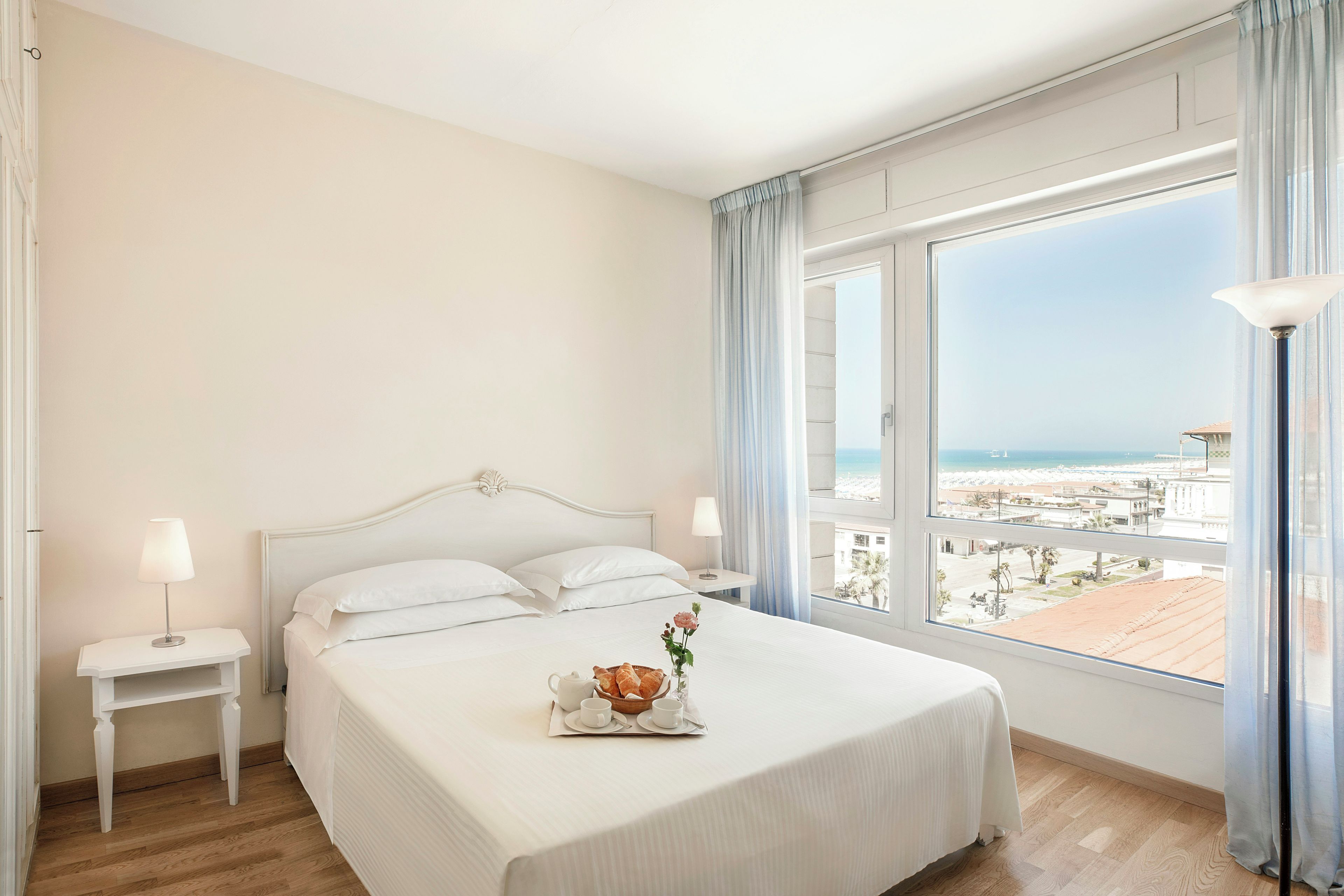 A hotel room with a bed, a bedside table, a lamp, a tray with breakfast, and a view of the beach.