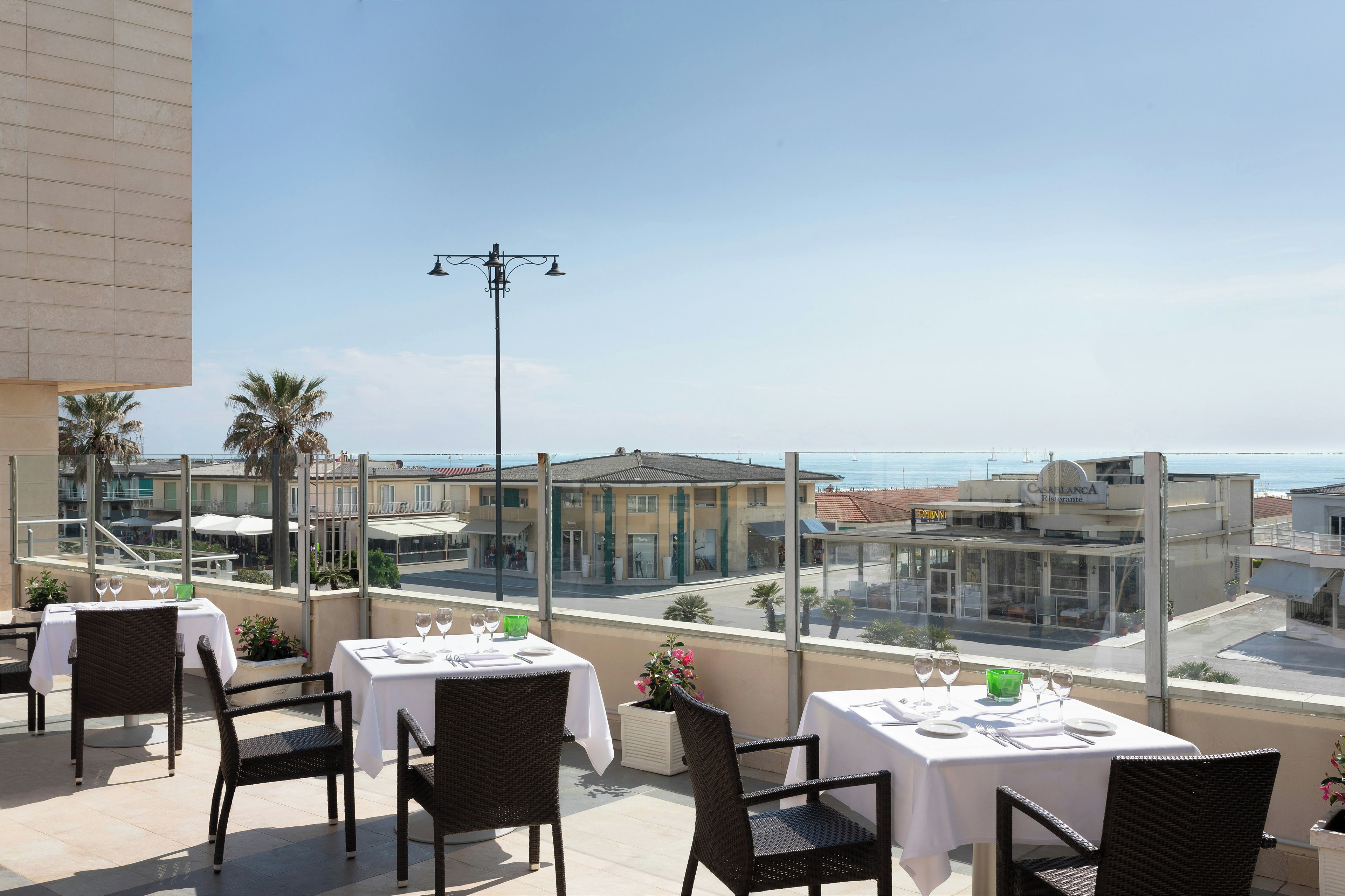 A terrace with tables set for dining, overlooking a coastal area with buildings and a clear sky.