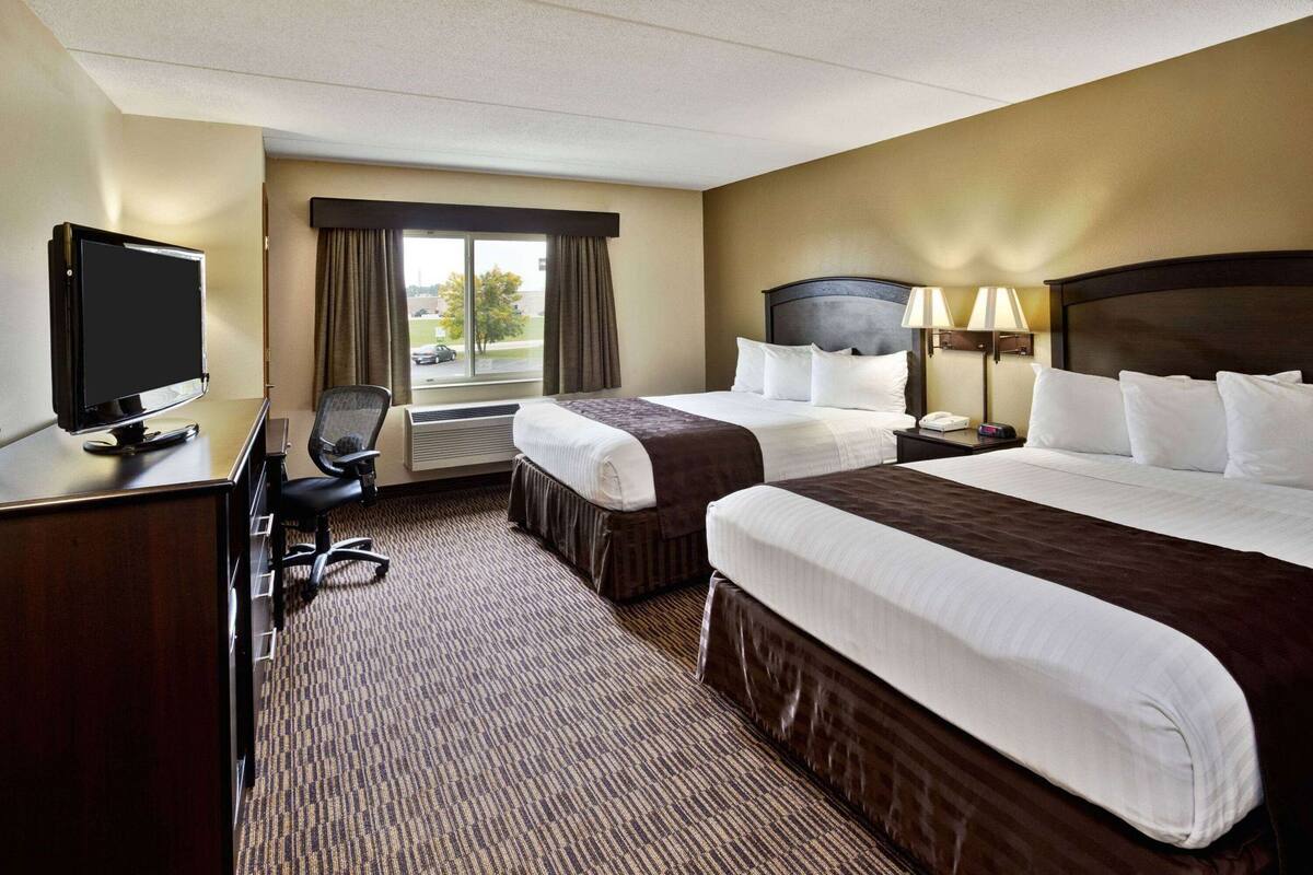 A hotel room with two beds, a desk with a TV, a chair, a lamp, and a window with curtains.