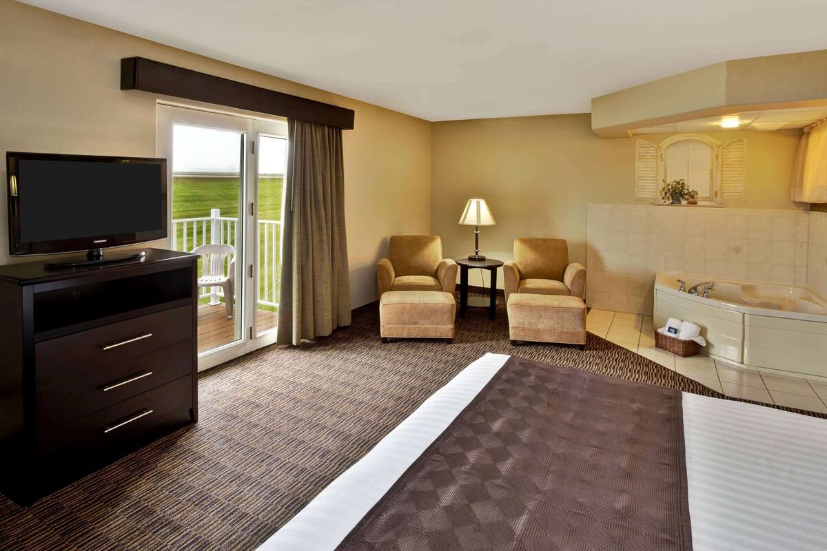 A hotel room with a bed, a TV, two armchairs, a small table with a lamp, a dresser, a balcony with curtains, and a bathroom with a bathtub.