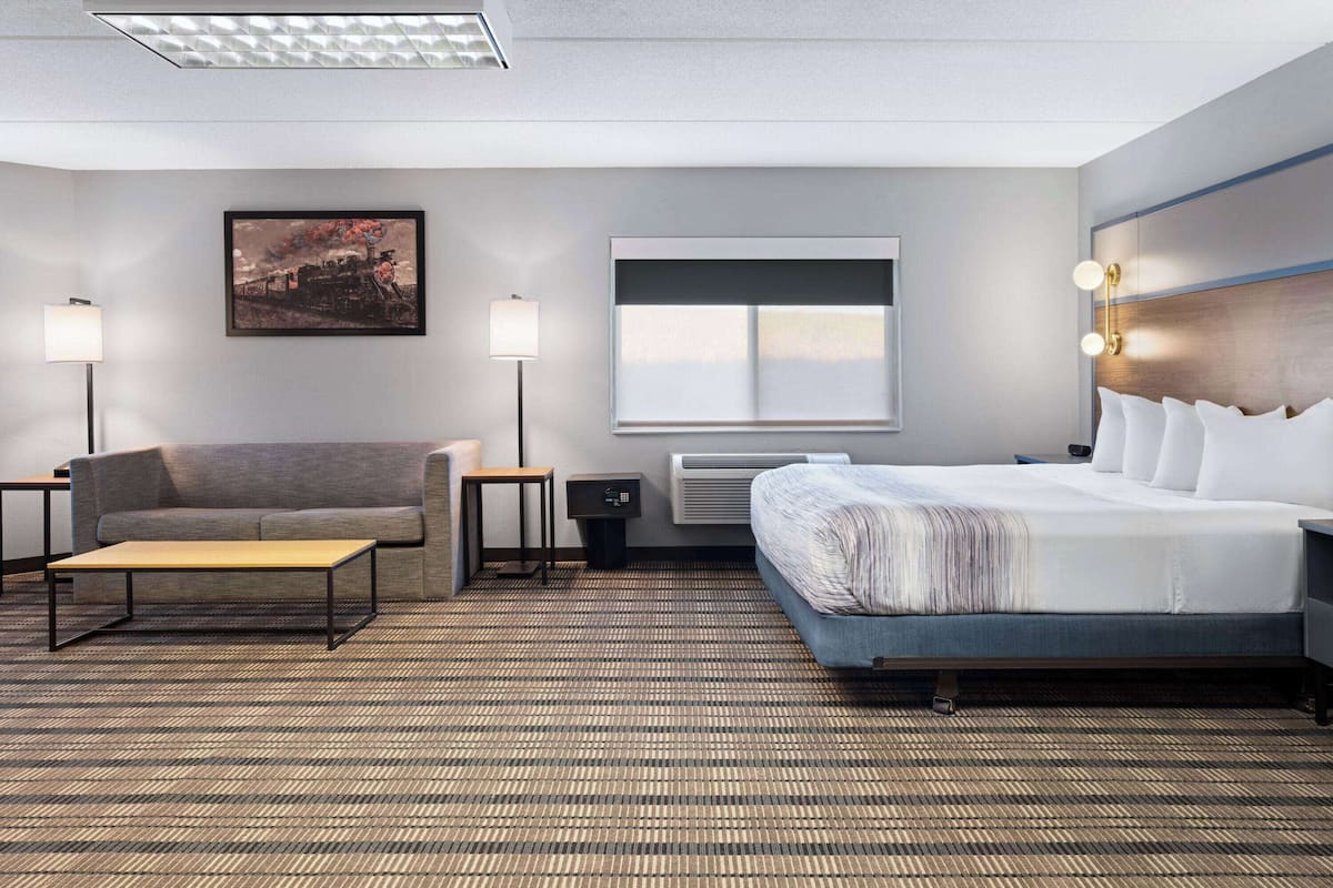 A hotel room with a bed, a sofa, a coffee table, two lamps, a painting on the wall, and a window.
