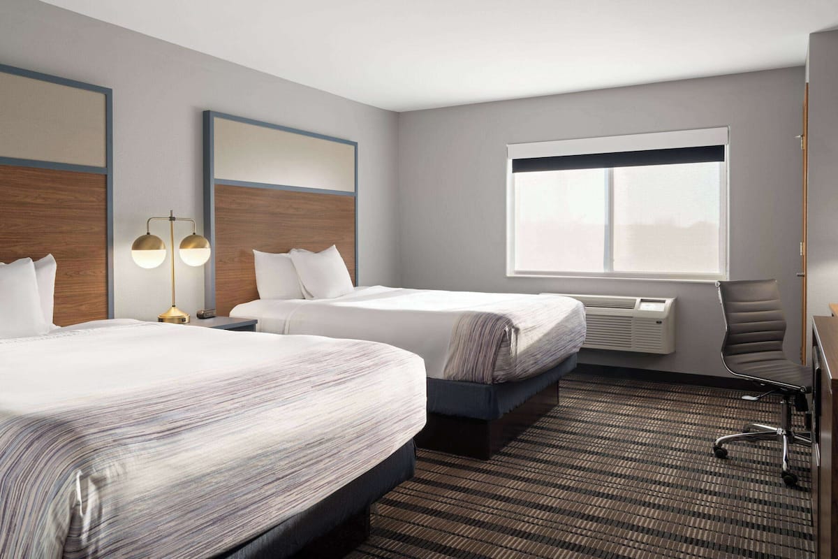 A hotel room with two beds, a desk, a chair, a lamp, and a large window.