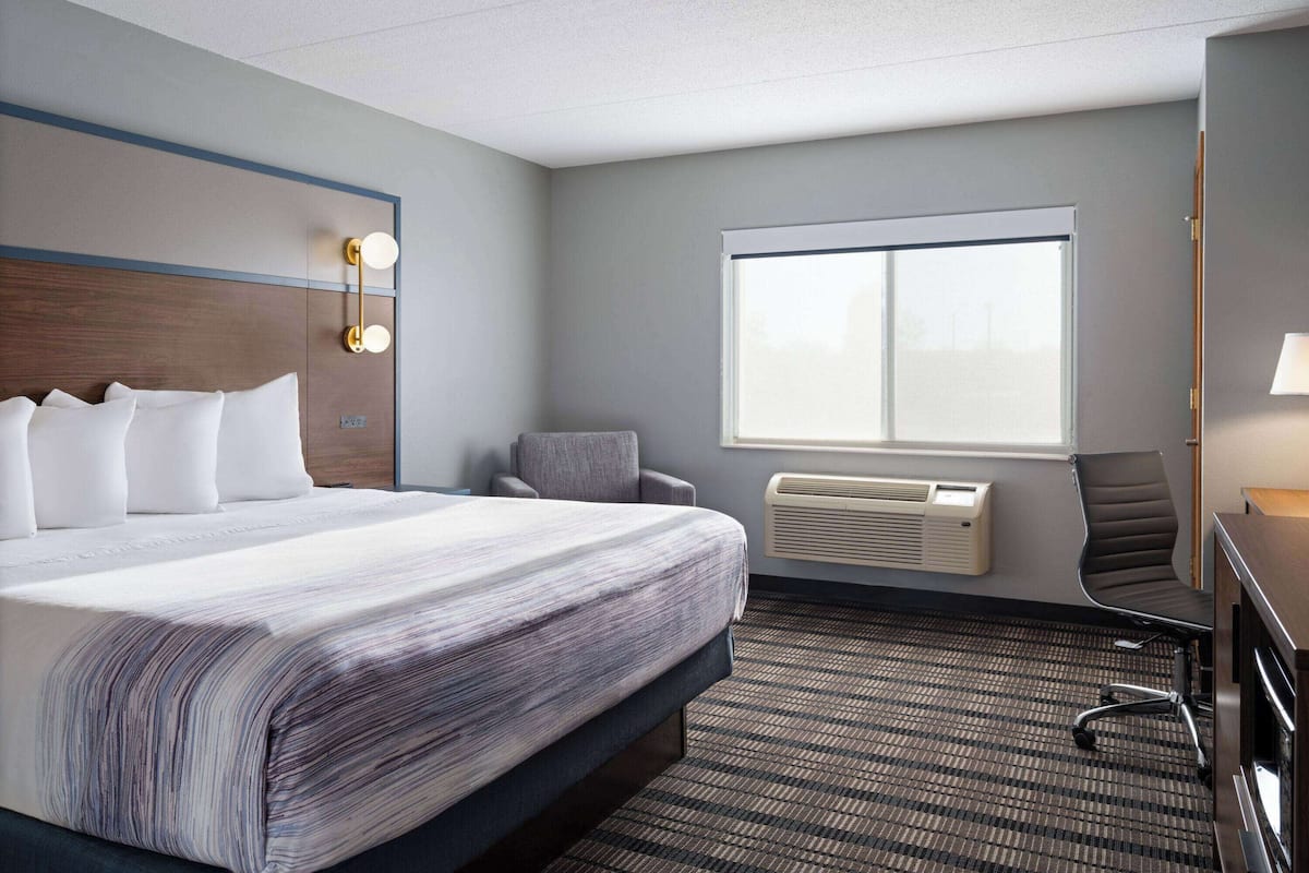 A hotel room with a large bed, a desk with a lamp, a chair, and a window with blinds.
