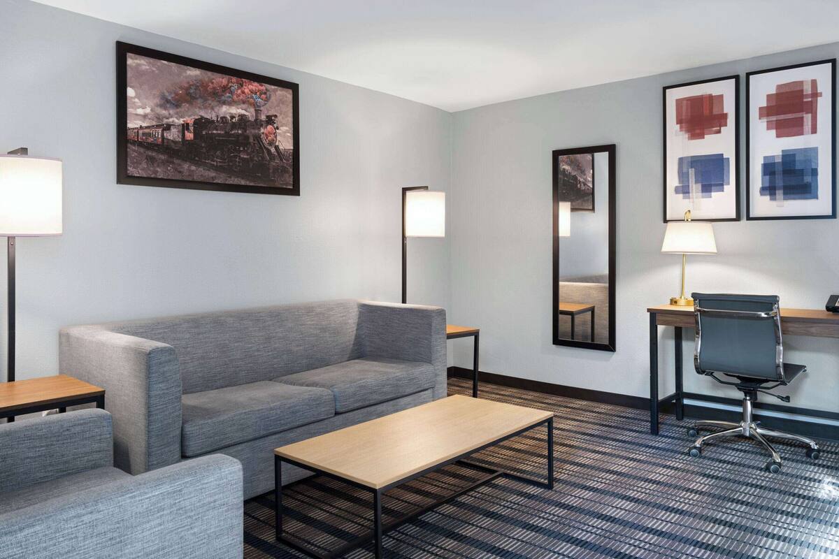 A hotel room with a gray sofa, a wooden coffee table, a desk with a chair, and artwork on the walls.