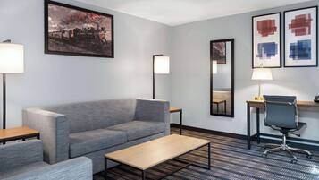 A hotel room with a gray sofa, a wooden coffee table, a desk with a chair, and artwork on the walls.