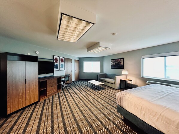 A hotel room with a bed, a desk with a chair, a sofa, a TV, and a window. - AmericInn by Wyndham Beaver Dam (Beaver Dam)